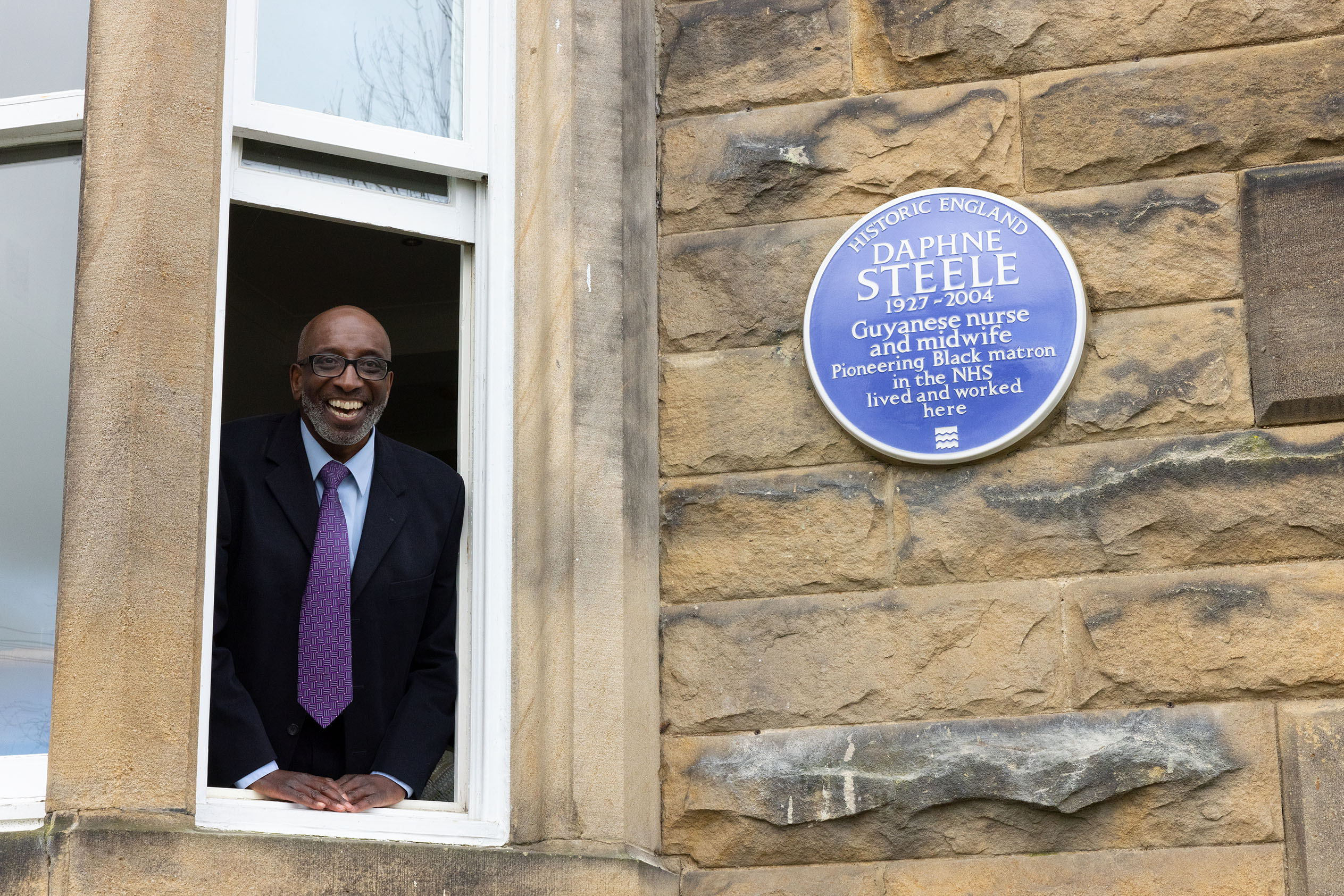 A man in a suit and tie smiling at camera while stood at an open window next to a heritage blue plaque