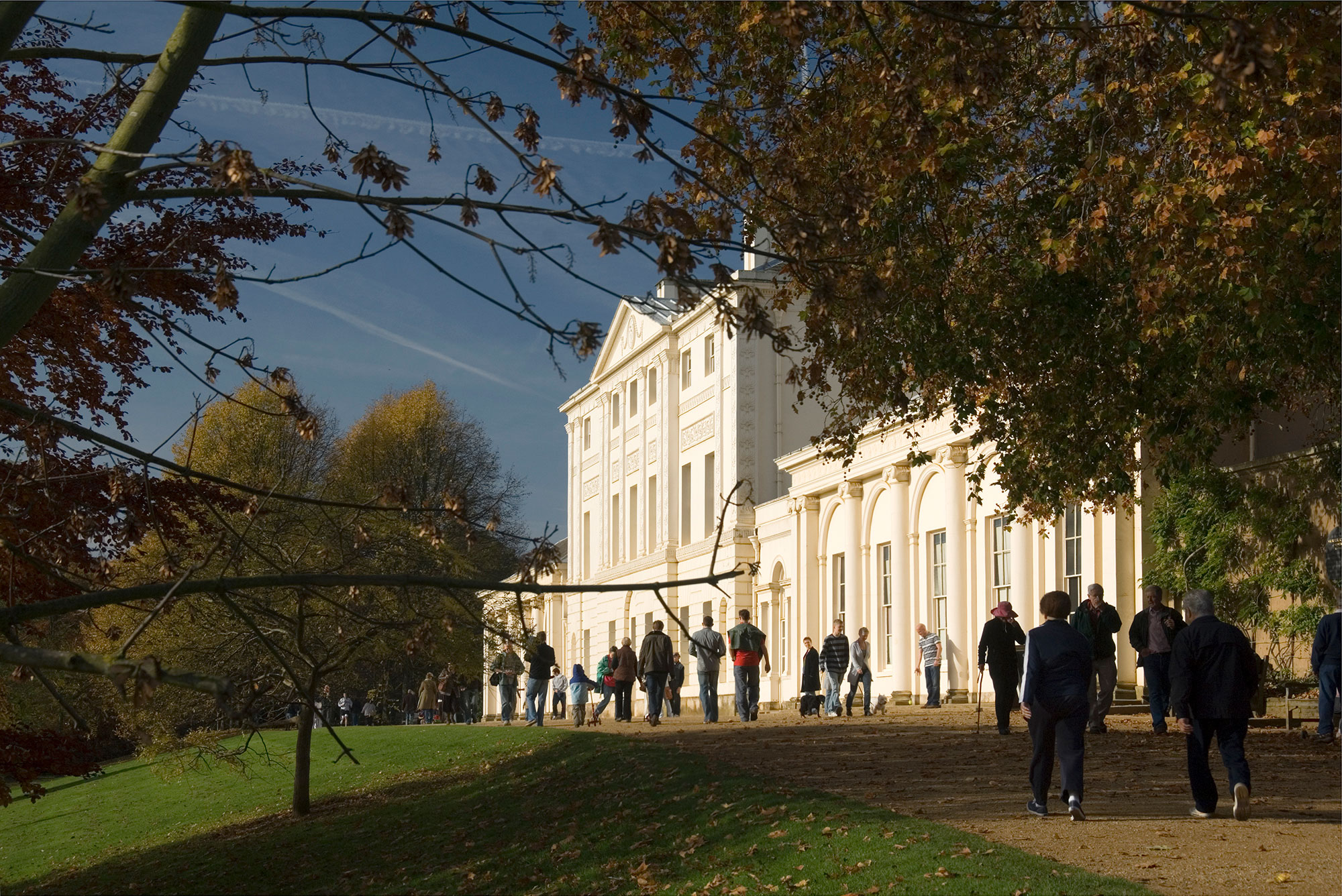 Photo of the a sunlit front of Kenwood House with people walking along the path in front