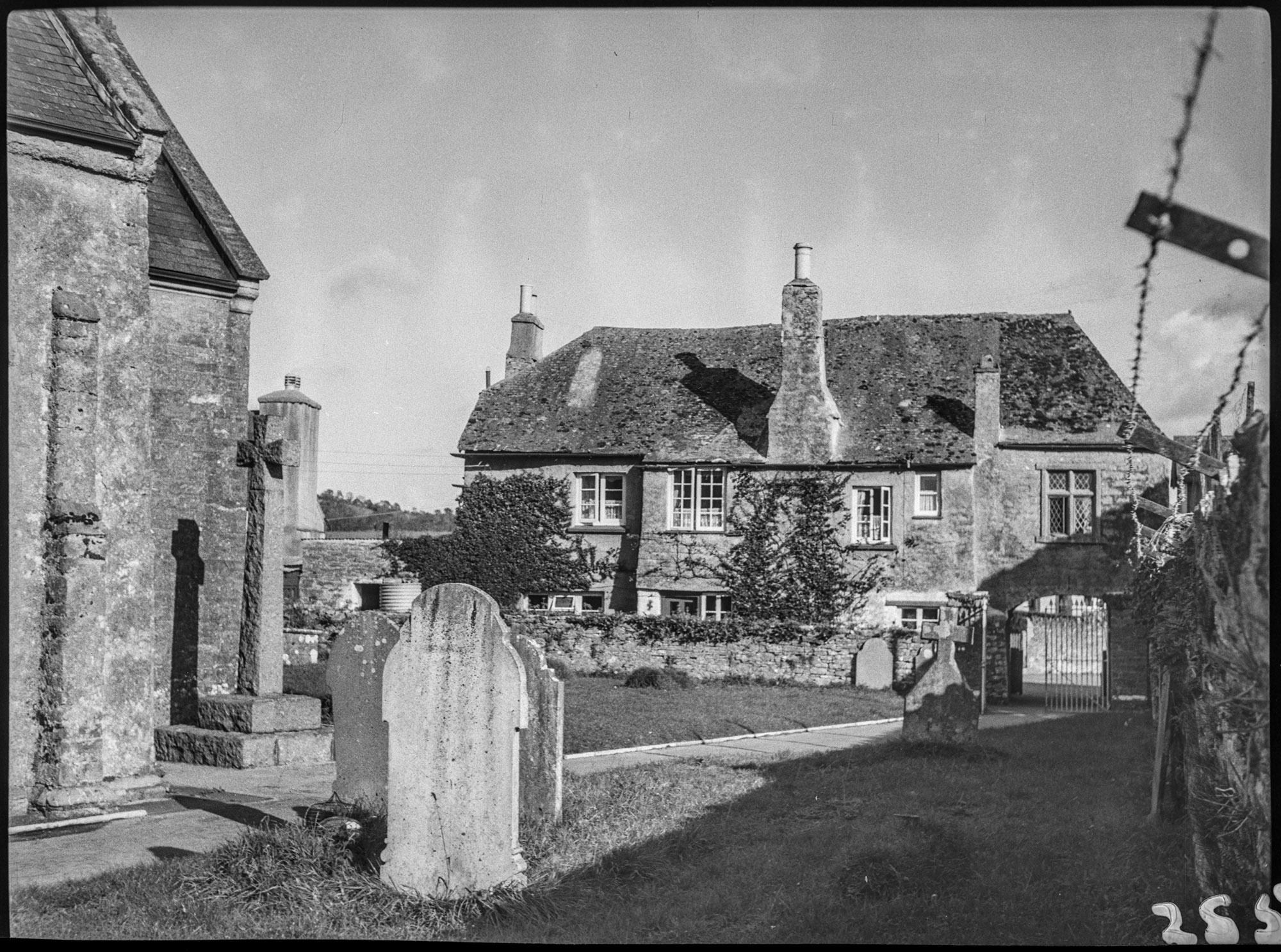 Black and white photograph showing part of a church and churchyard in the foreground and a two-storey stone building in the background. To the right of the image is a stone wall topped with barbed wire.