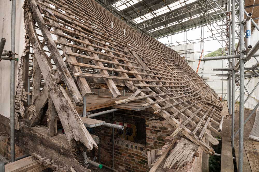 A photograph of a historic timber roof structure enclosed under protective scaffolding. The timbers are in a poor state of repair.
