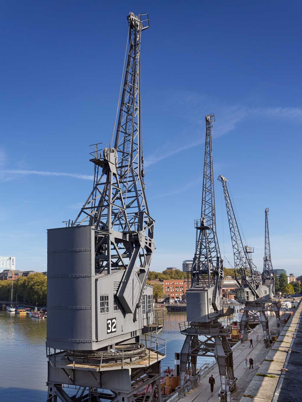 A photograph of four large, grey, metal cranes on a dockside with the river beyond.