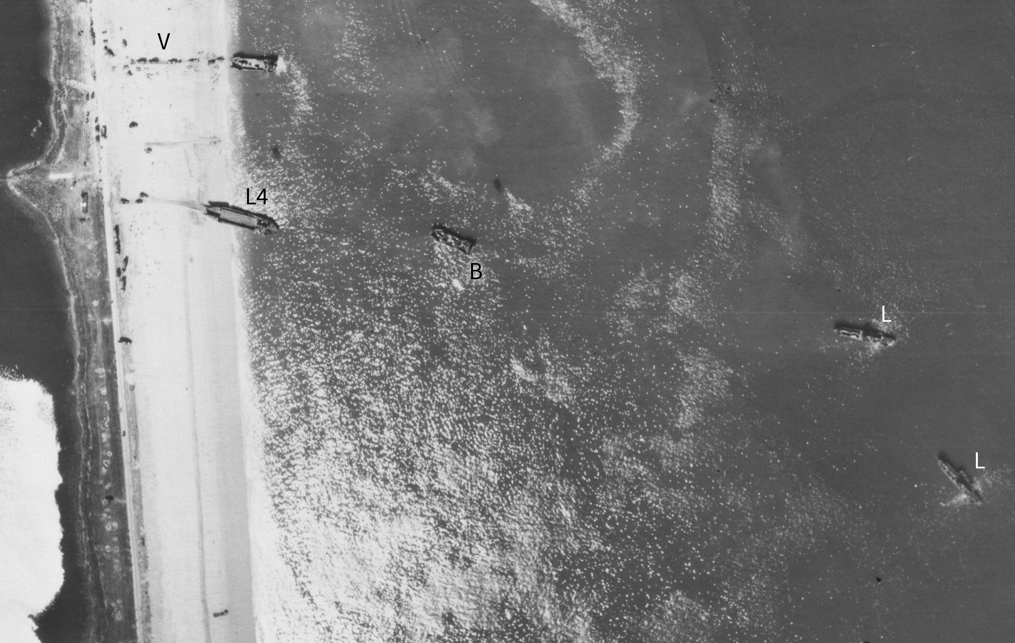 Detail from a black and white vertical aerial photograph, showing a narrow strip of beach between an inland pool of water and the sea. Two landing craft are stationary at the edge of the beach. A line of vehicles are on the beach next to the open doors of one of the landing craft. Vehicle tracks on the sand lead from the second landing craft to vehicles further up the beach. Other vehicles are dotted on the beach. At sea are three other vessels. One appears to have a barrage balloon tethered to it. Sunlight illuminates the water towards the bottom-left of the image. The letters and numbers 'V', 'L4', 'B' and 'L' applied to the image mark locations.