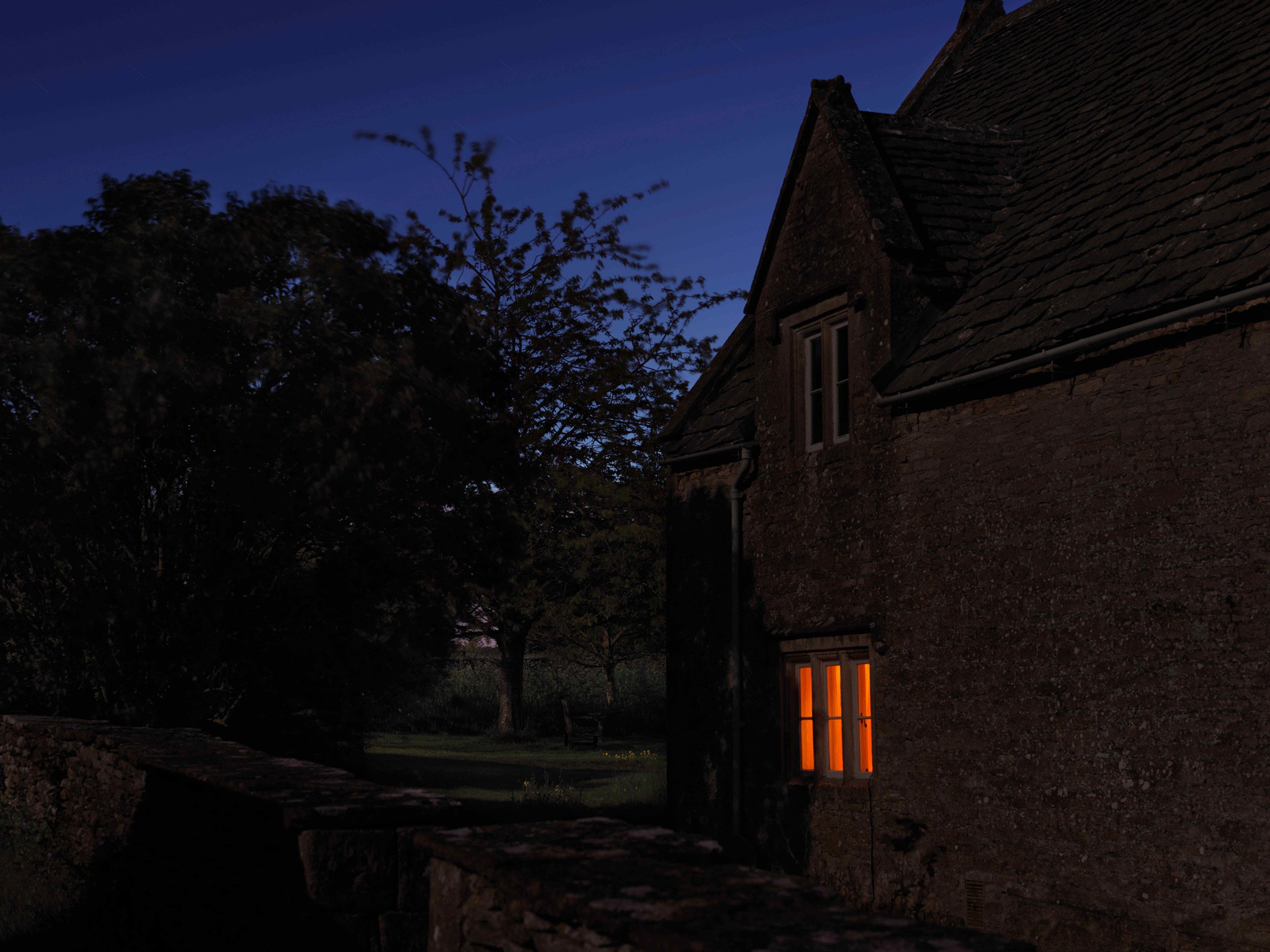 Orange light shines through a window at twilight. The home is made of Cotswold stone.