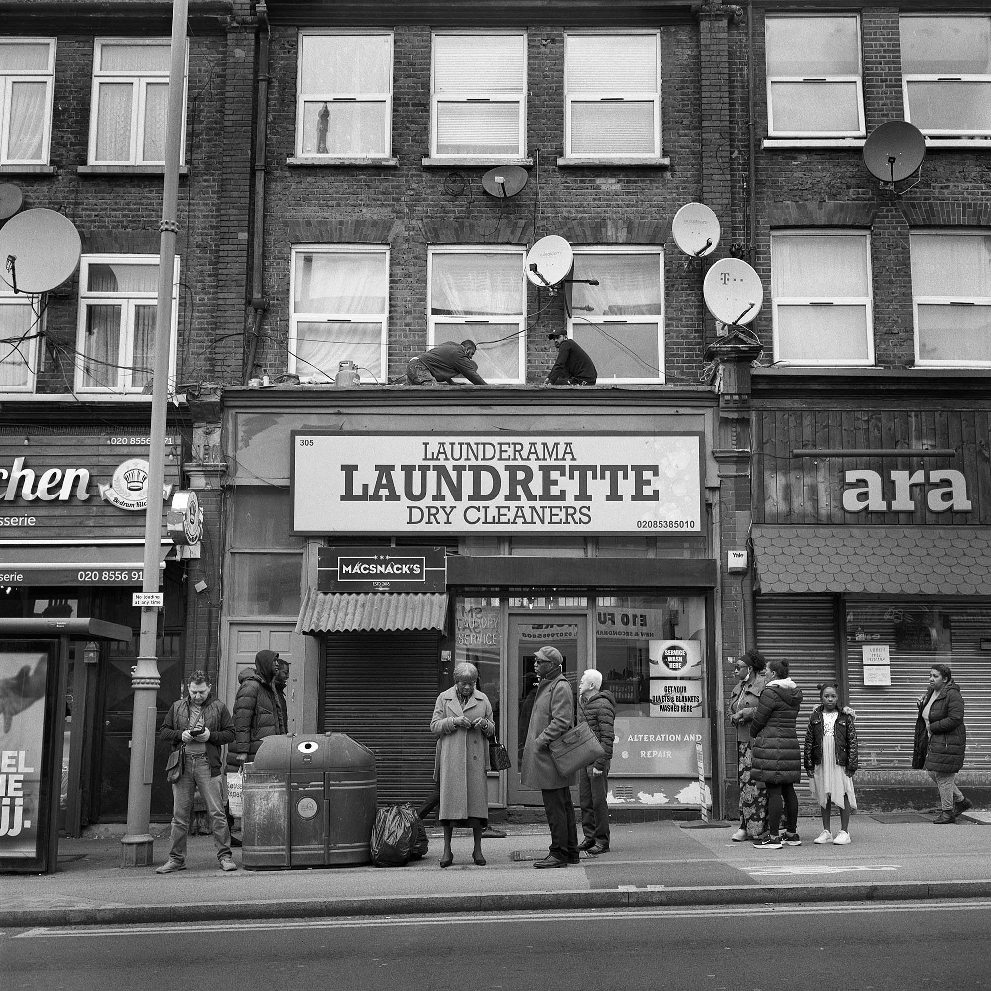 Black and white image of people waiting at a bus stop and in the background are people working on the roof above the launderette sign.