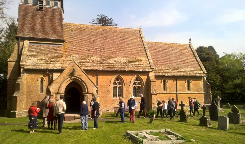 A photograph of people gathering outside a small, honey-coloured church set in a well-kept graveyard.