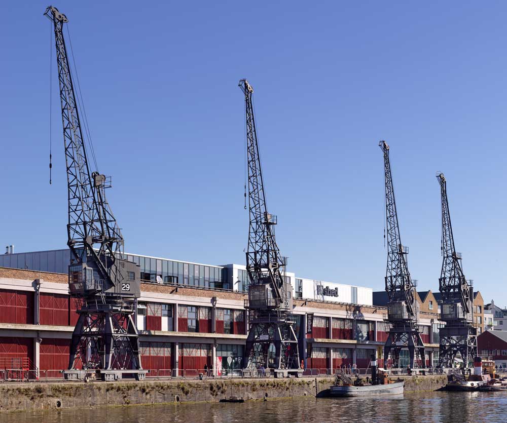 A photograph of four large, grey, 20th century cranes on a wharf with a long industrial building behind them.