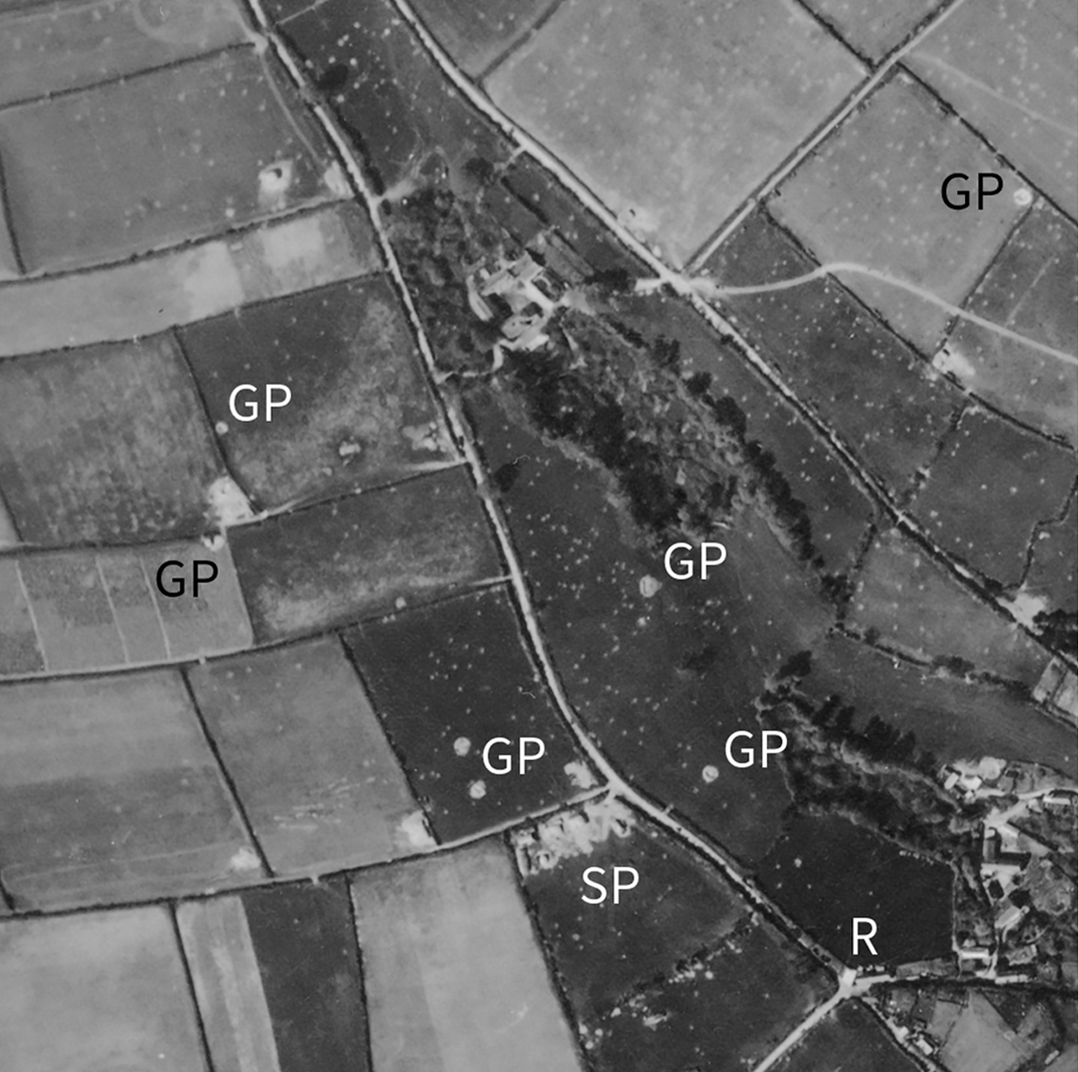 Detail from a black and white vertical aerial photograph, showing a patchwork landscape of fields, hedges, roads and tracks. In the centre of the image and in the bottom-right corner are buildings. The fields are pockmarked with shell holes. The letters 'R', 'SP' and 'GP' applied to the image mark locations