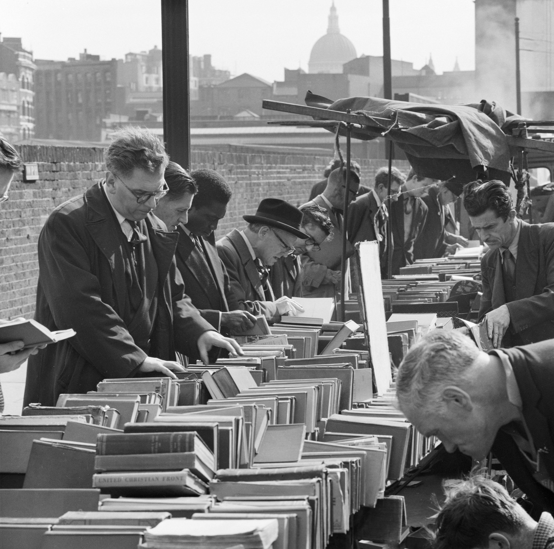 A black and white photo showing men in suits and overcoats browsing books on an open-air bookstall. The stall extends diagonally from the bottom-left. Behind a row of browsers is a brick wall. Amongst the background buildings, the dome of St Paul's Cathedral is visible.