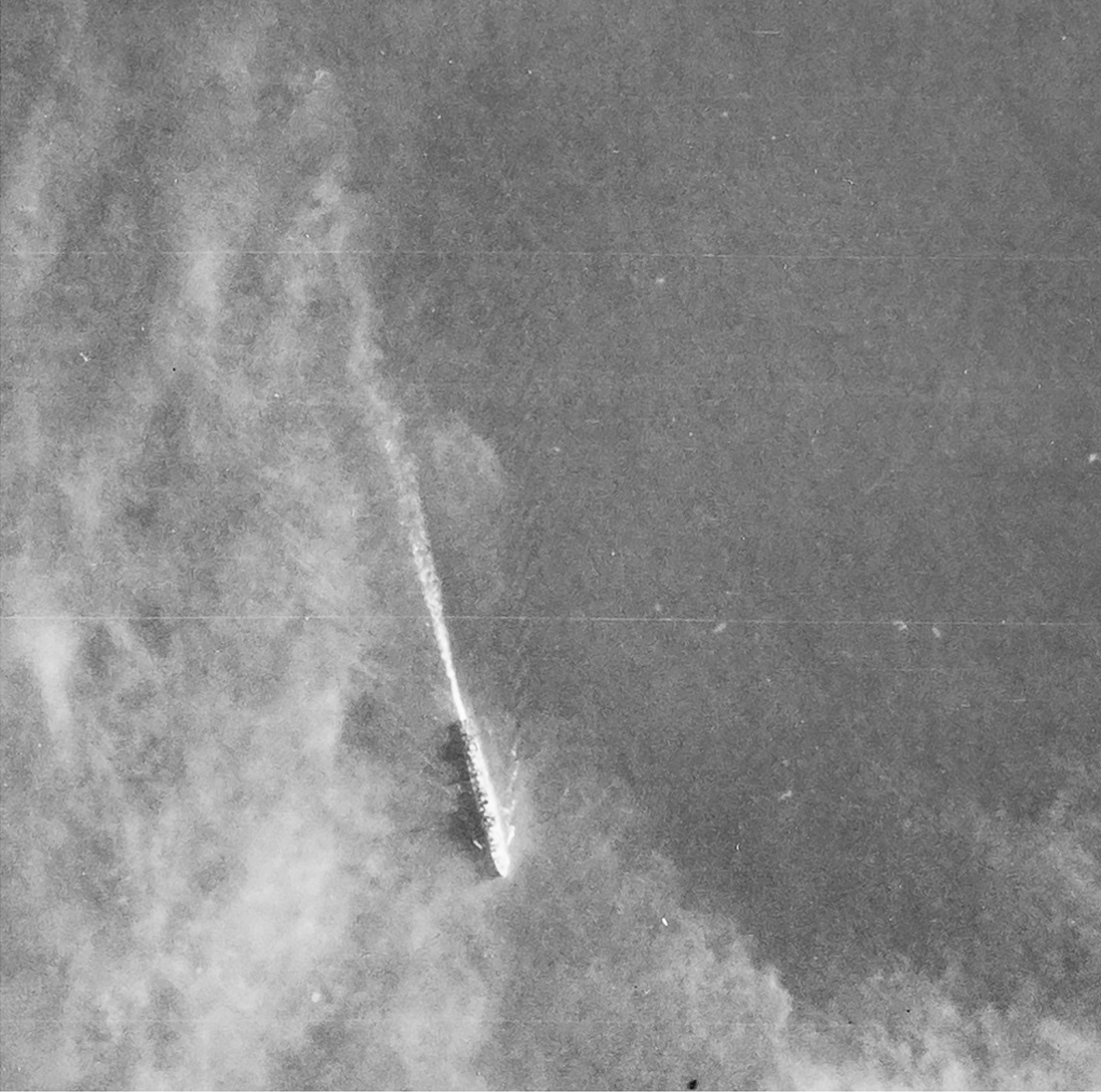 Detail from a black and white vertical aerial photograph, showing a warship at sea. A straight line of wake trails behind the ship. Above the ship, wafts of smoke partly obscure the view.