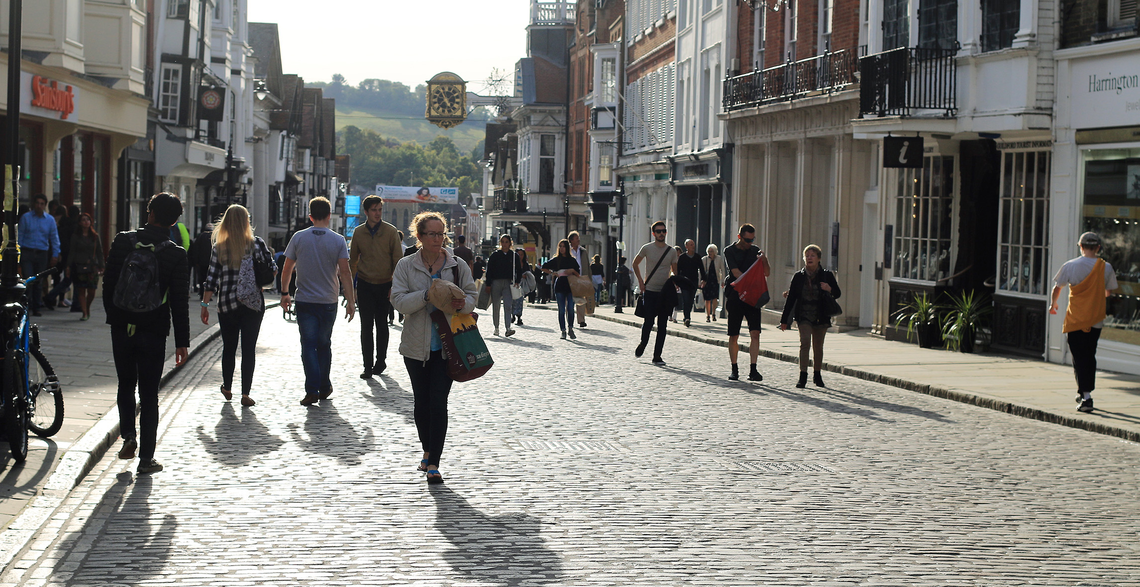 Guildford high street on a busy and sunny day. Pedestrians are walking both on the pavements and down the middle of the cobbled street.