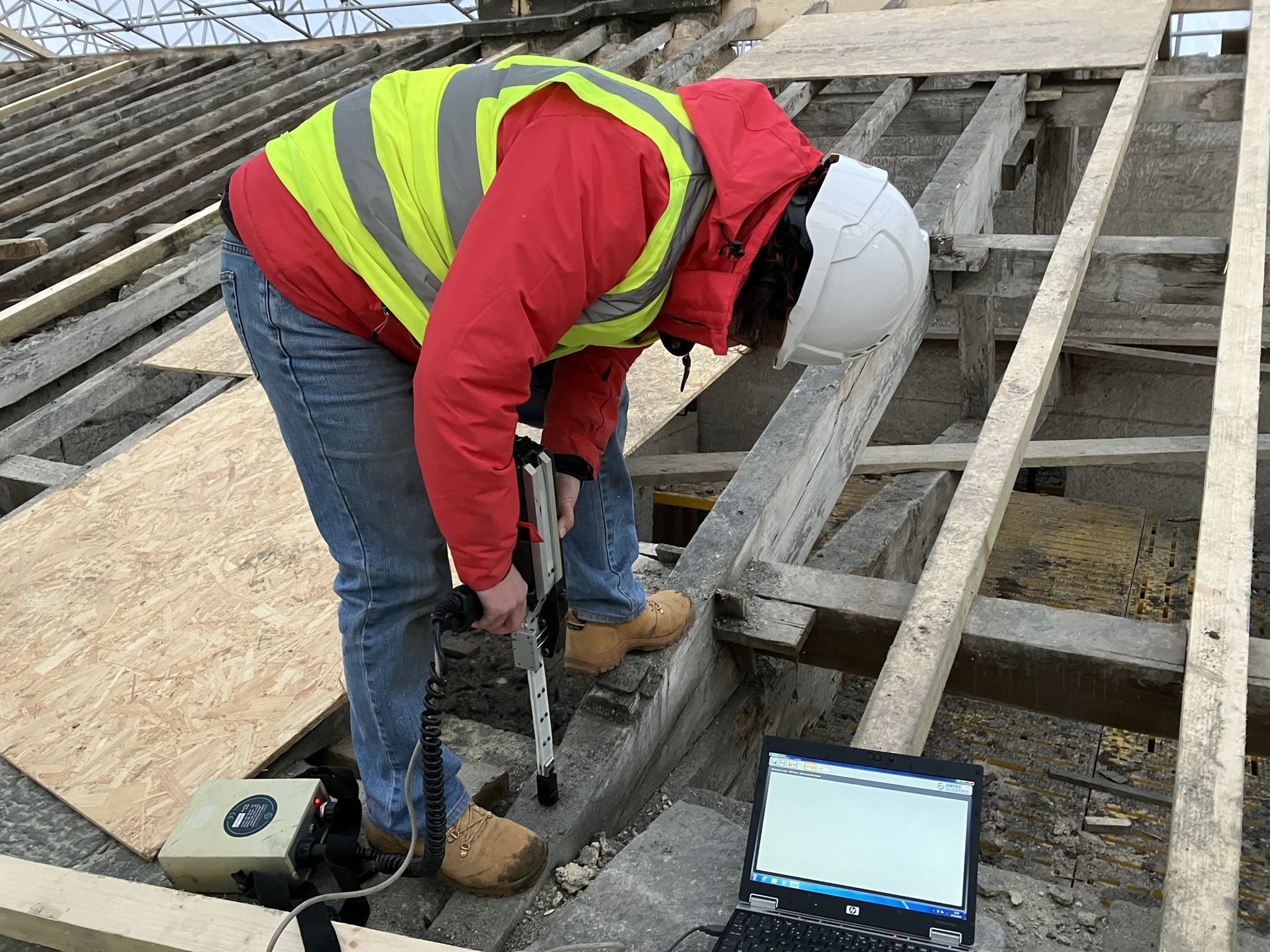 Photograph showing dark-haired woman in red coat, high vis and hard hat using a decay detection drill on Belsay Hall roof trusses. 