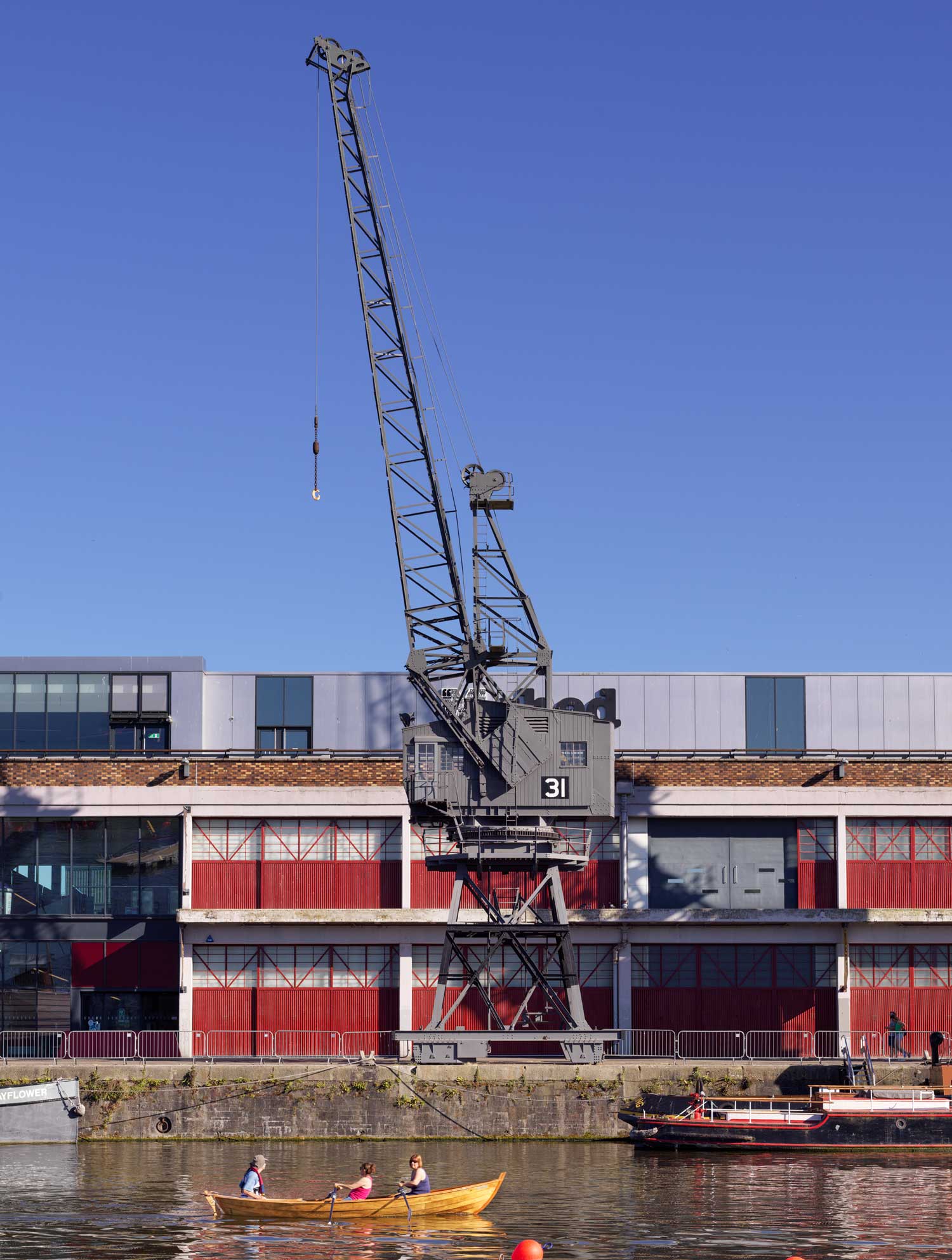 A photograph looking across a river to a large metal crane on the dockside, in front of an industrial building.