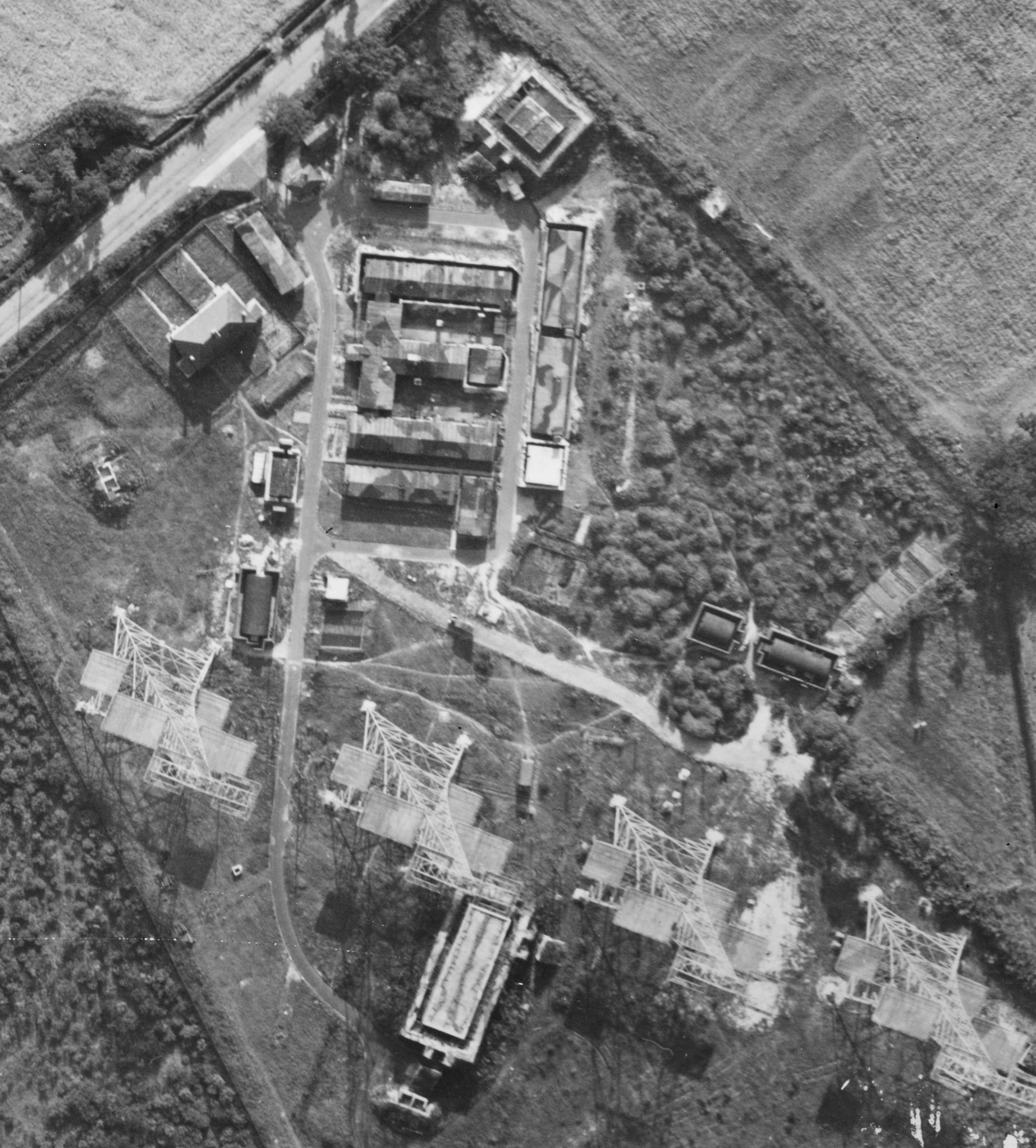 A black and white vertical aerial photograph of four lattice towers adjacent to buildings with camouflaged roofs and a pair of semi-detached houses. A stretch of road passes diagonally in the top-left corner.