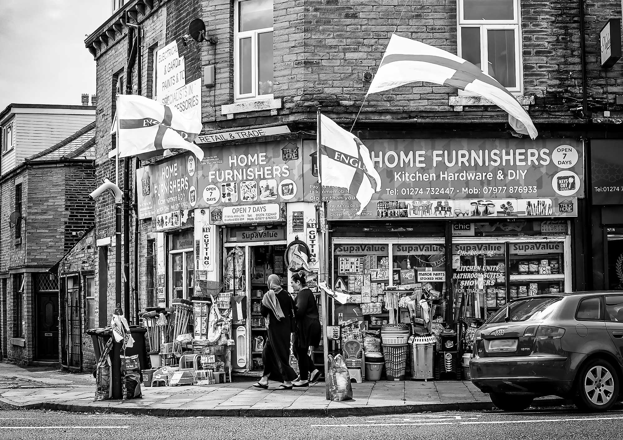 A black and white photo of a shop on the corner of two streets. The shop windows are full of display items and further items are set out on the pavement. Two people walk on the pavement towards the shop entrance. Shop sign reads: 'Home Furnishers'. Three flags of St George fly from flag poles attached to the shop. 