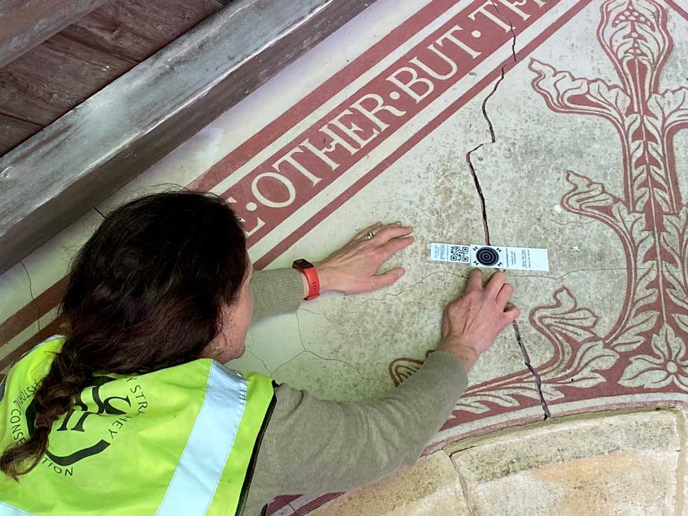 A photograph of a woman fixing a small monitoring device to a crack in a richly decorated interior church wall.