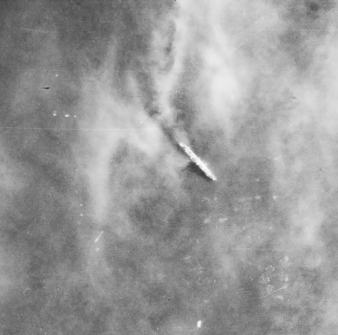 Detail from a black and white vertical aerial photograph, showing a warship at sea. Above the ship, wafts of smoke partly obscure the view.