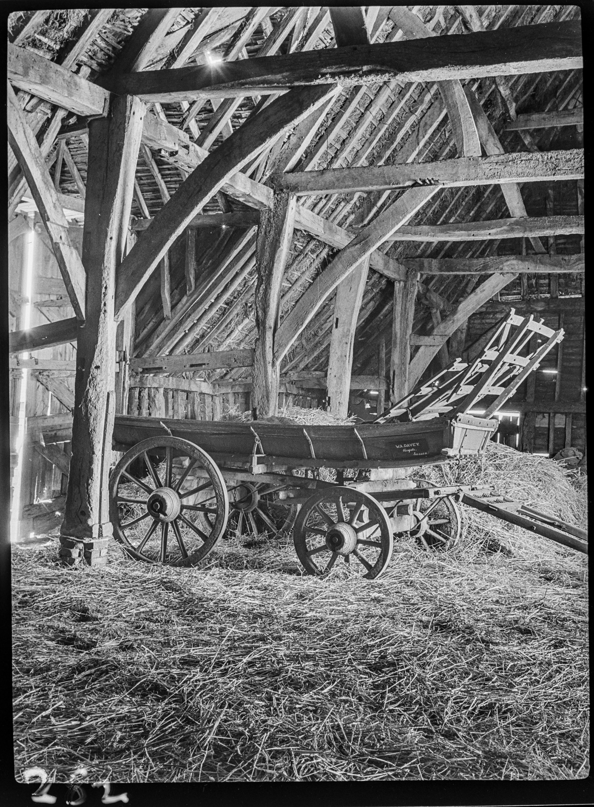 Black and white photograph showing part of the interior of a large agricultural barn constructed with timber trusses. Straw is strewn on the floor of the barn. In the centre of the photograph is a static, wooden cart. A sign on the cart reads: 'W.A. Davey Rogate Sussex'.