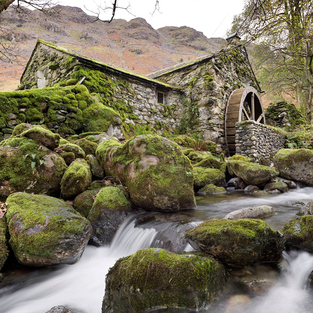A stone-built water mill on a rocky stream in front of a tall hill.