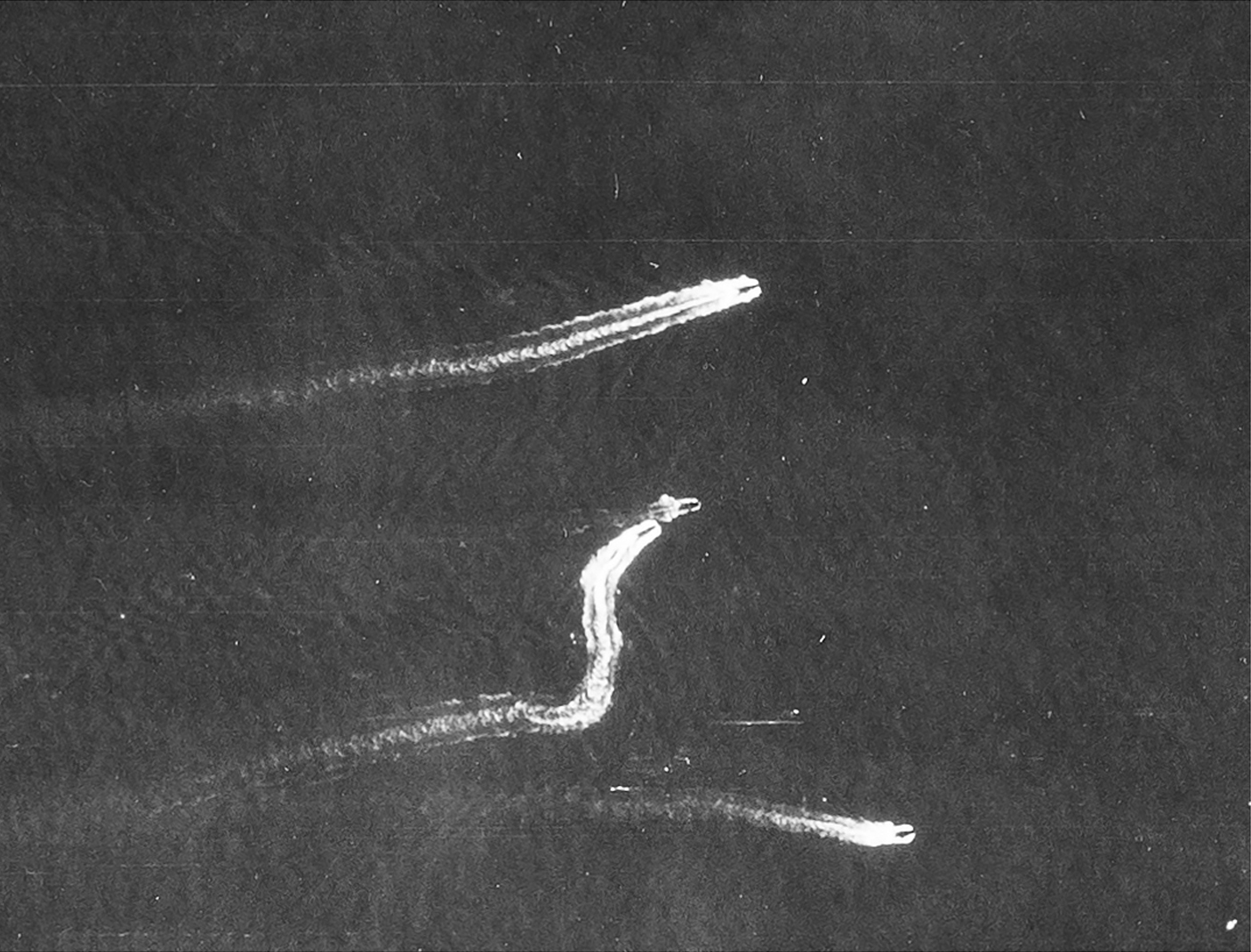 Detail from a black and white vertical aerial photograph, showing four landing craft at sea. Two vessels sail towards the right edge of the image. Between them, two others make an 'S'-shaped wake as they manoeuvre in the water.