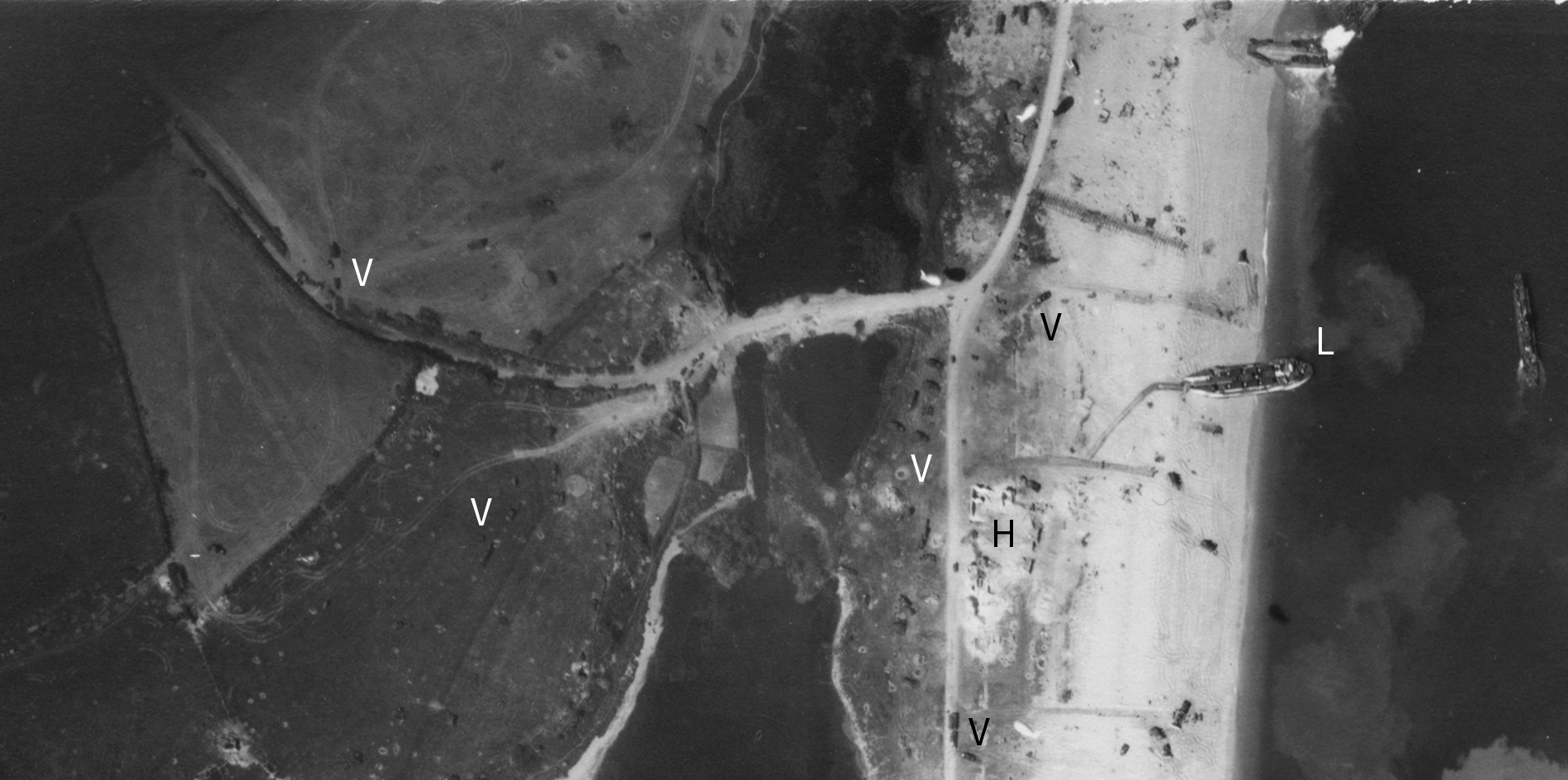Detail from a black and white vertical aerial photograph, showing a narrow strip of beach between an inland pool of water and the sea. Towards the centre-top of the image is a bridge spanning a narrow section of the pool of water. A beachside road crosses the bridge and continues inland. Two large landing craft are stationary at the edge of the beach. Tracks lead from them. Other vehicles are on the beach. Barrage balloons float above the landing area. The letters 'L', 'H' and 'V' applied to the image mark locations.