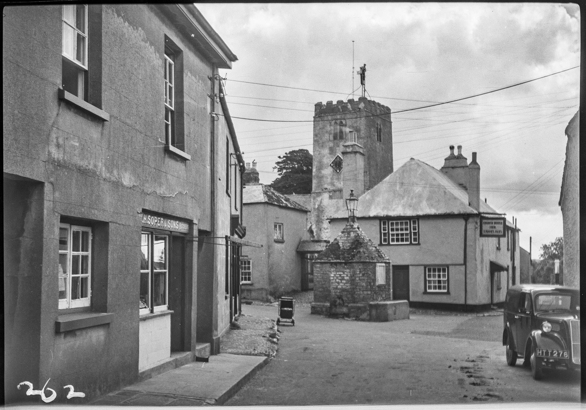 Black and white photograph showing a street view. To the left is a row of shops and houses. In the centre is a squat, stone building with a pyramidical roof. Adjacent to this and next to the pavement is a pram. In the background, a square church tower rises above two buildings, one of which is a public house. In the right foreground, part of a car can be seen parked adjacent to the wall of a building.