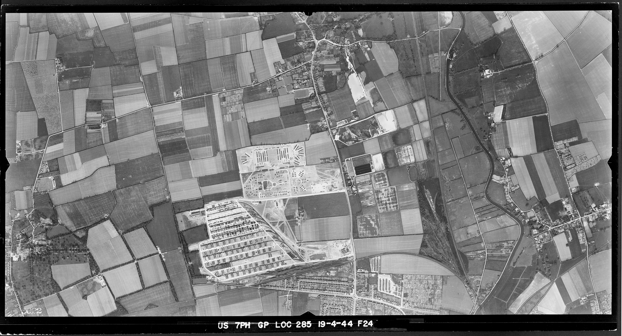 A black and white vertical aerial photograph showing a storage depot and camp bounded by roads and a patchwork of fields.
