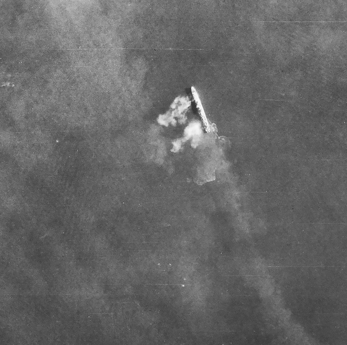 Detail from a black and white vertical aerial photograph, showing a waship at sea. Puffs of smoke to the left of the ship suggest that its guns are firing. Above the ship, wafts of smoke partly obscure the view.