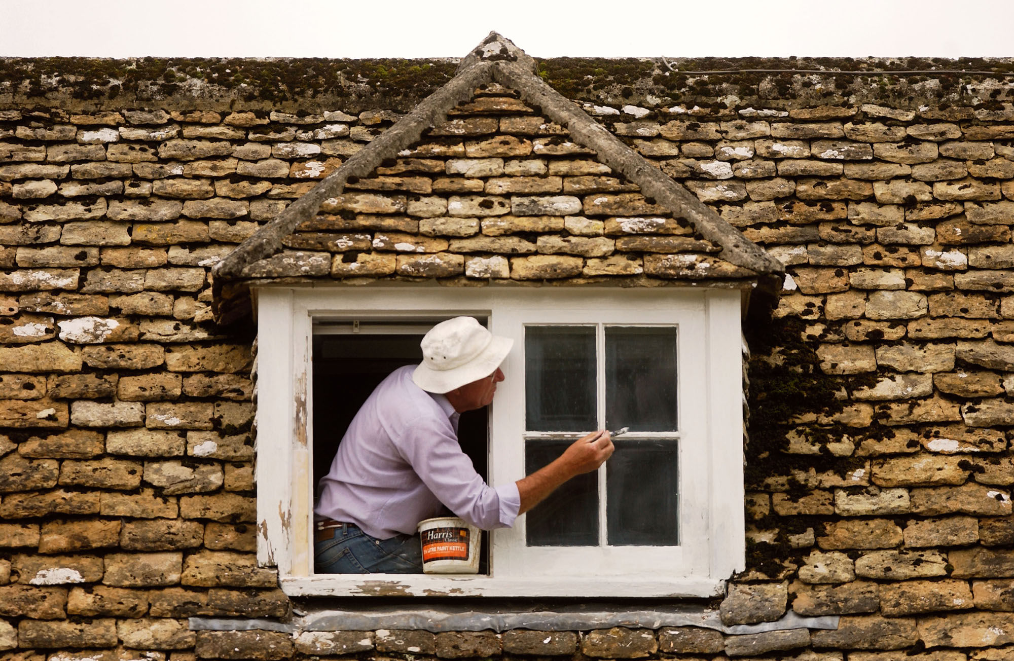 A man leaning out of a dormer window of an old cottage to paint it. 