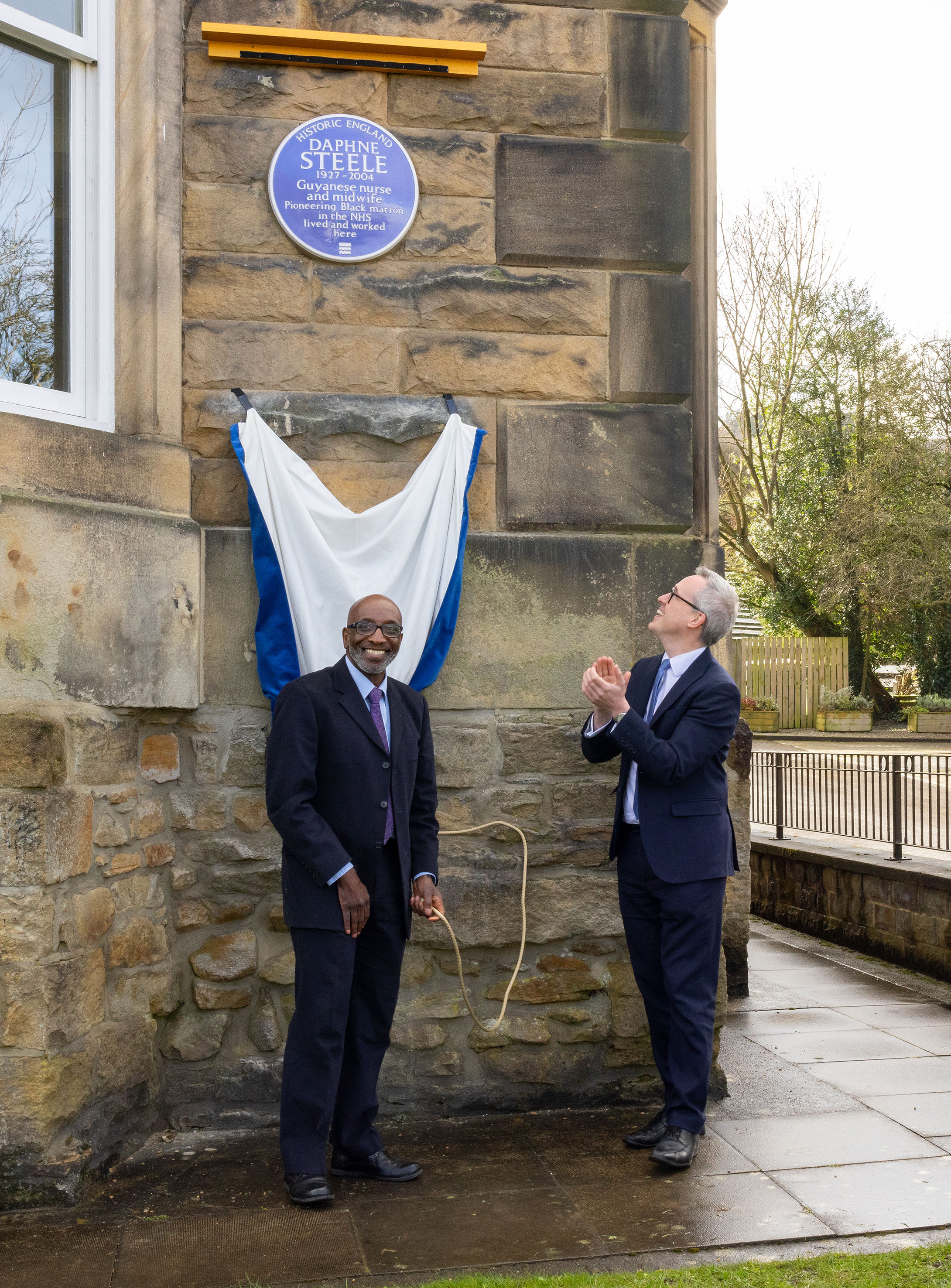 Two smiling men in suits standing underneath a heritage blue plaque in the moments after its unveiling