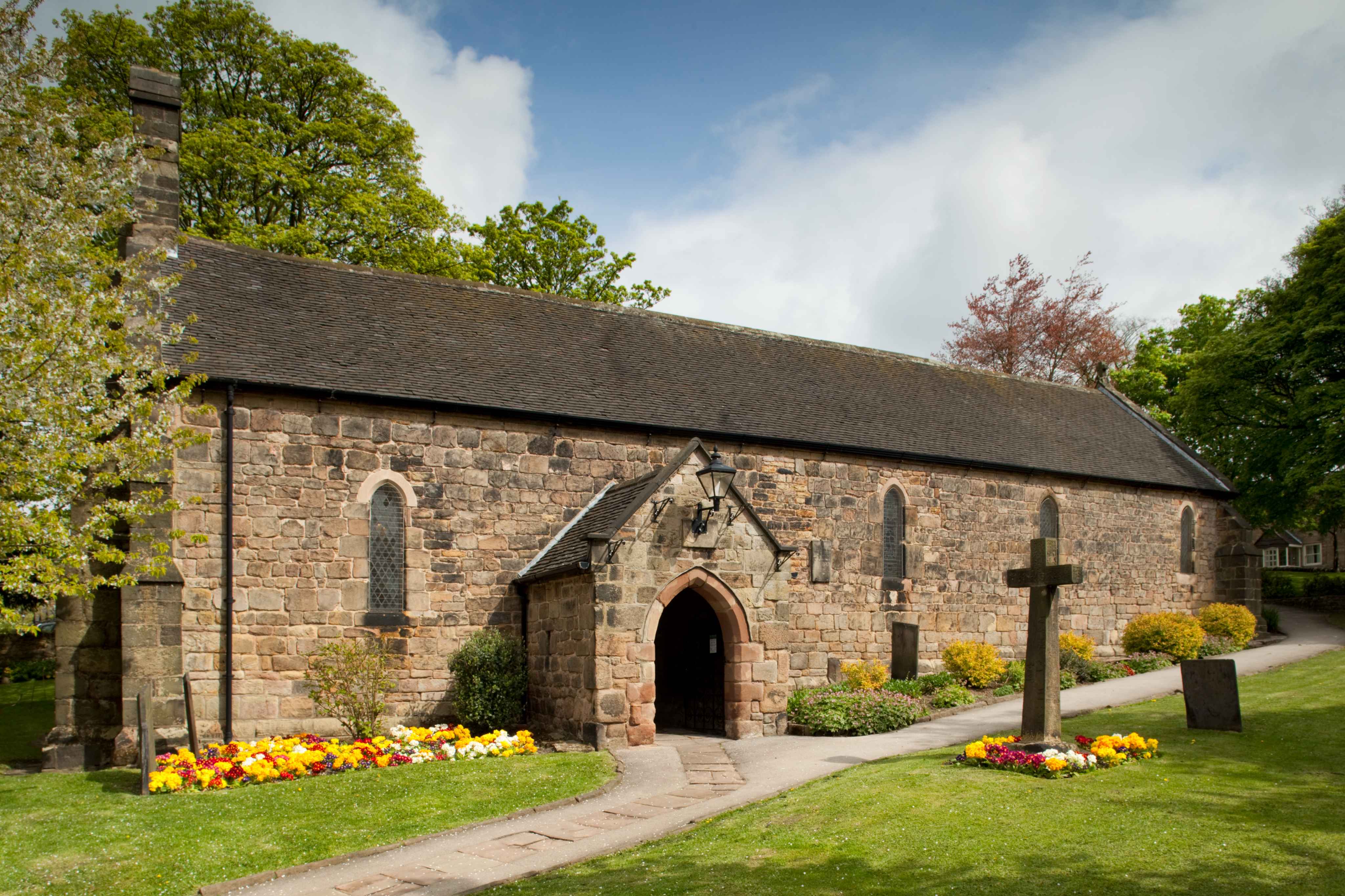Image of the exterior of a small church with a stone cross outside and neat flowerbeds.