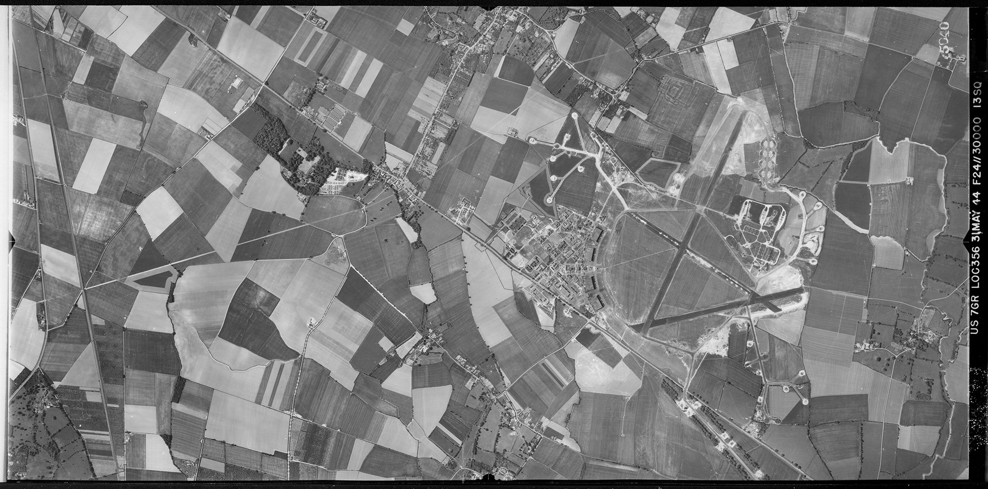 A black and white vertical aerial photograph of an airfield with three runways set within a landscape formed of a patchwork of fields. 