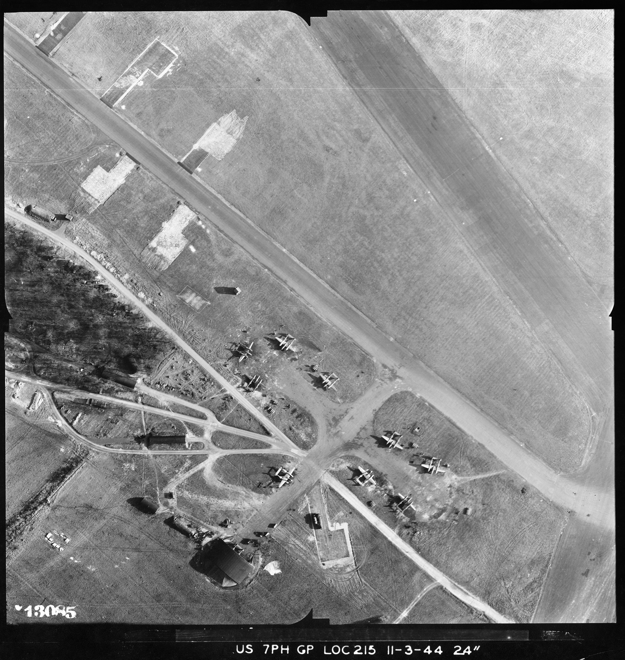 A black and white vertical aerial photograph showing part of an airfield. Running diagonally from top-left to bottom-right is a section of perimeter road. To the right of this is a section of runway. To the left is another road and a dispersal area with several parked aircraft and a small hangar. 