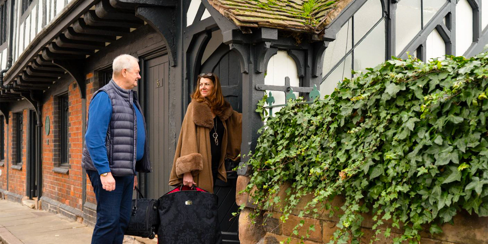 A couple with suitcases steps through the door of a half-timbered terraced house with a brick ground floor.