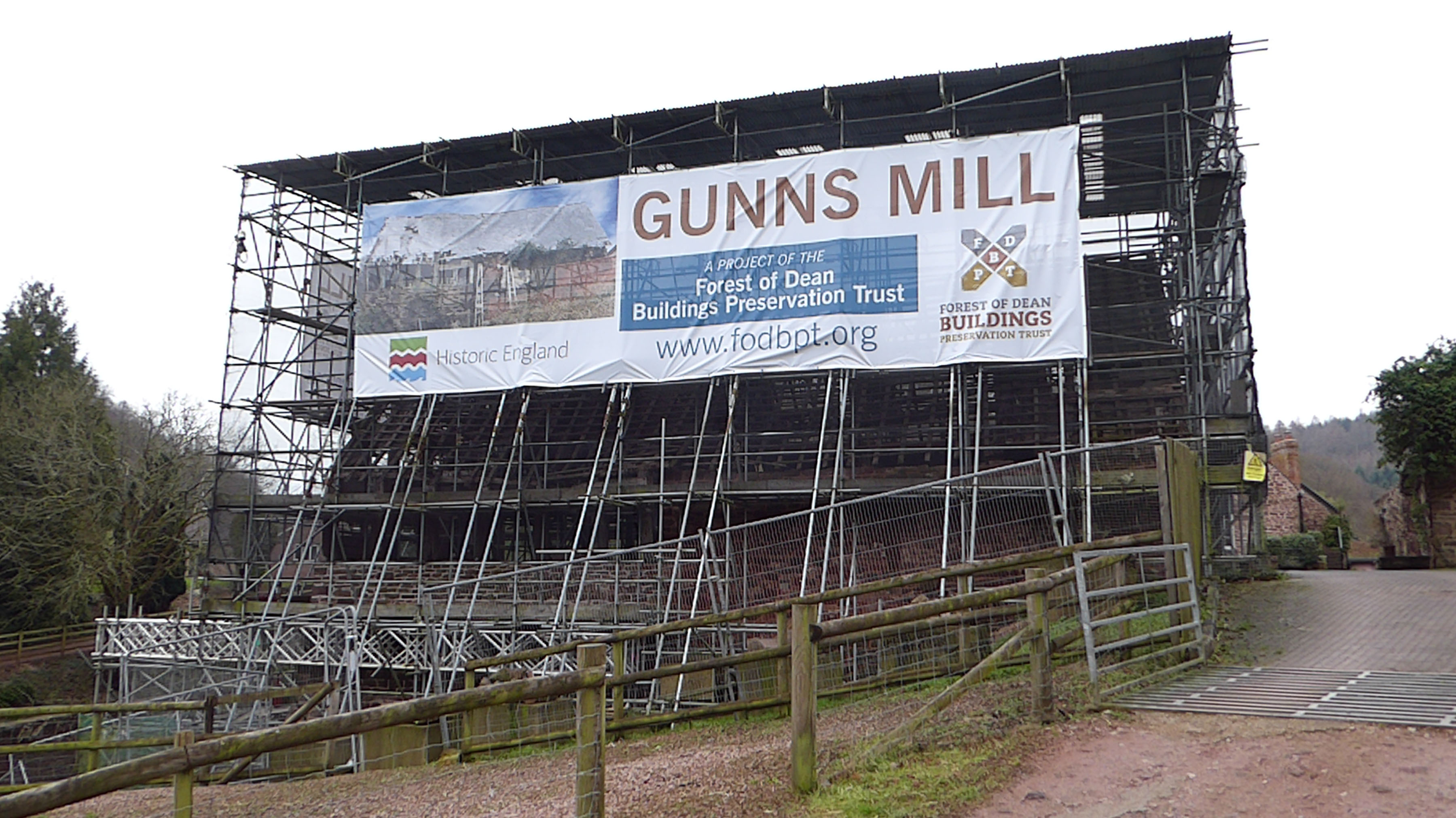 A photograph of a historic mill building entirely protected with scaffolding, showing a banner with the words 'Gunns Mill'.