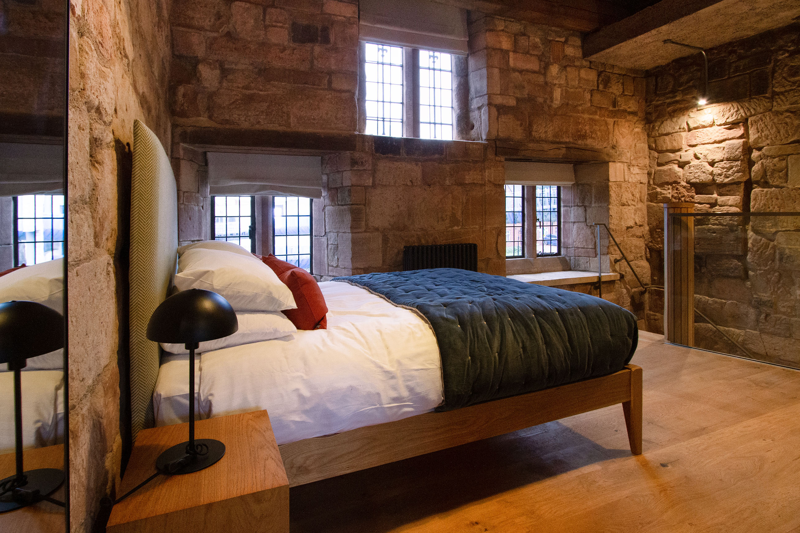 A modern double bed in a well-lit stone bedroom with three windows.