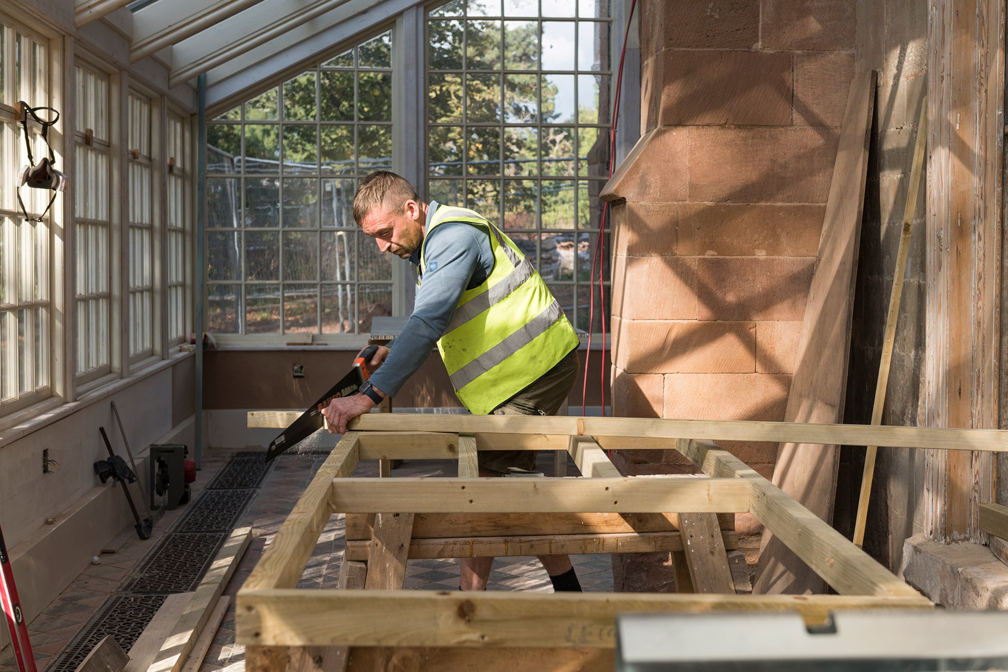 Man in a conservatory undergoing renovation. He is wearing a high-vis waistcoat and sawing a length of wood balanced on a frame made from the same wood.