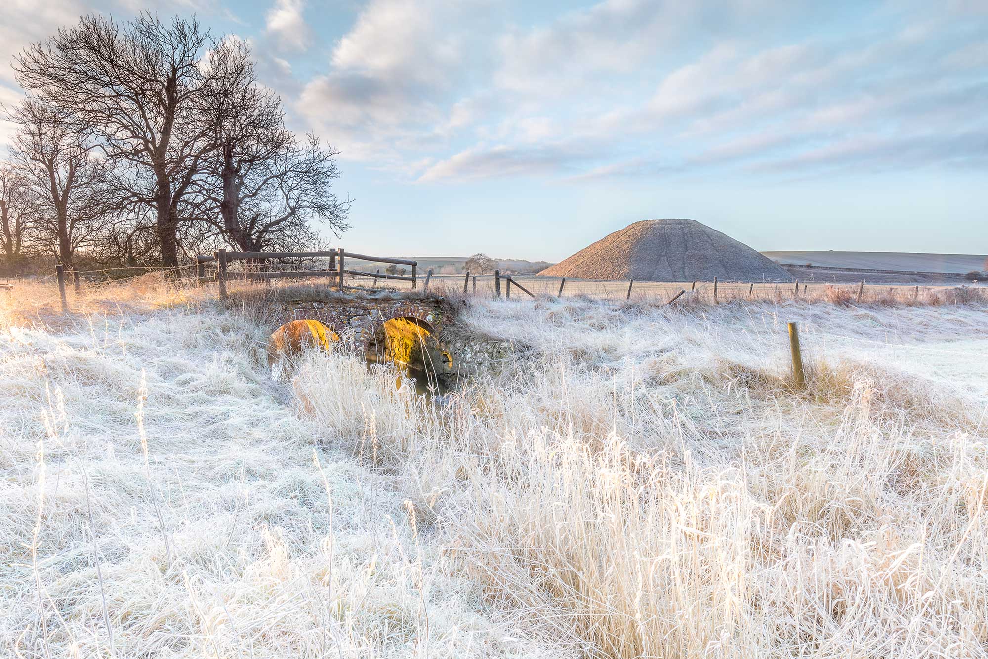 A photograph of a frosty countryside landscape, with a brick bridge in the foreground and a mound in the background.