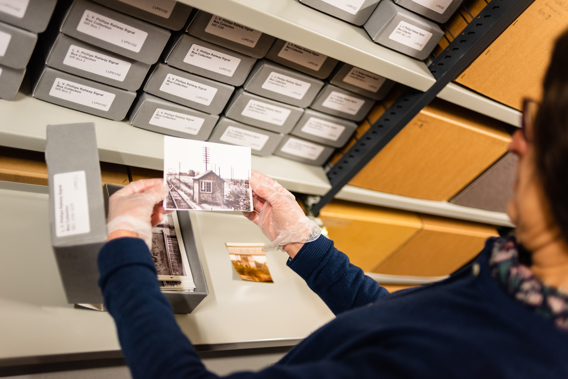A colour photograph being handled in the Historic England Archive store. The gloved hands of the member of staff carefully holds a photo of a small brick building next to a railway line. In the background are shelves with cardboard boxes. 