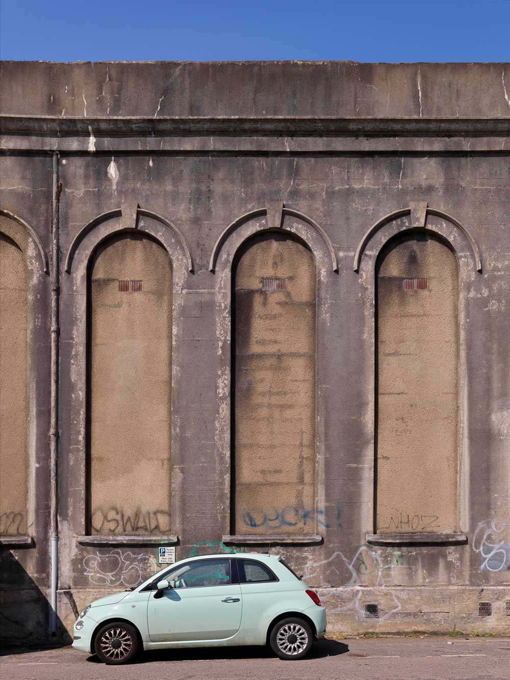 A photograph of a small car parked in front of a large concrete structure with a narrow blind arcade.
