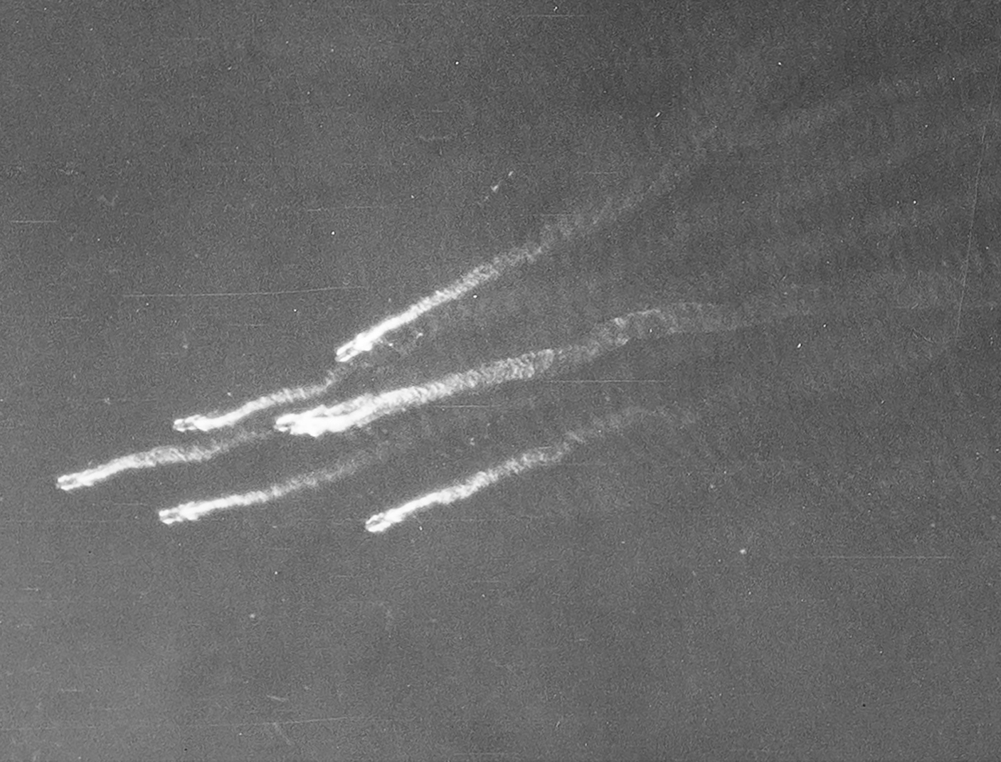 Detail from a black and white vertical aerial photograph, showing six landing craft at sea. The ships sail in an arrowhead formation towards the left edge of the image, trailing wake behind them.