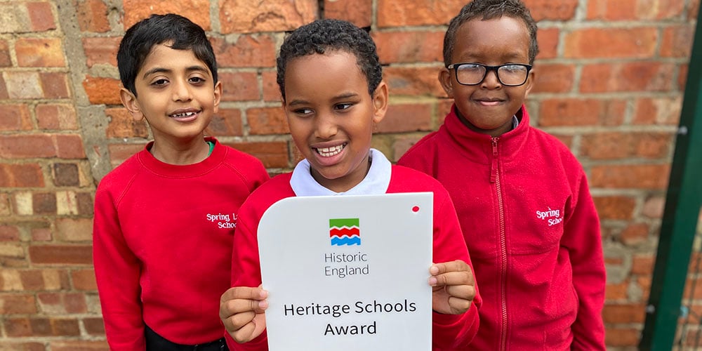 Three children in red school jumpers stand in front of a brick wall, holding up a plaque that reads "Historic England: Heritage Schools Award".
