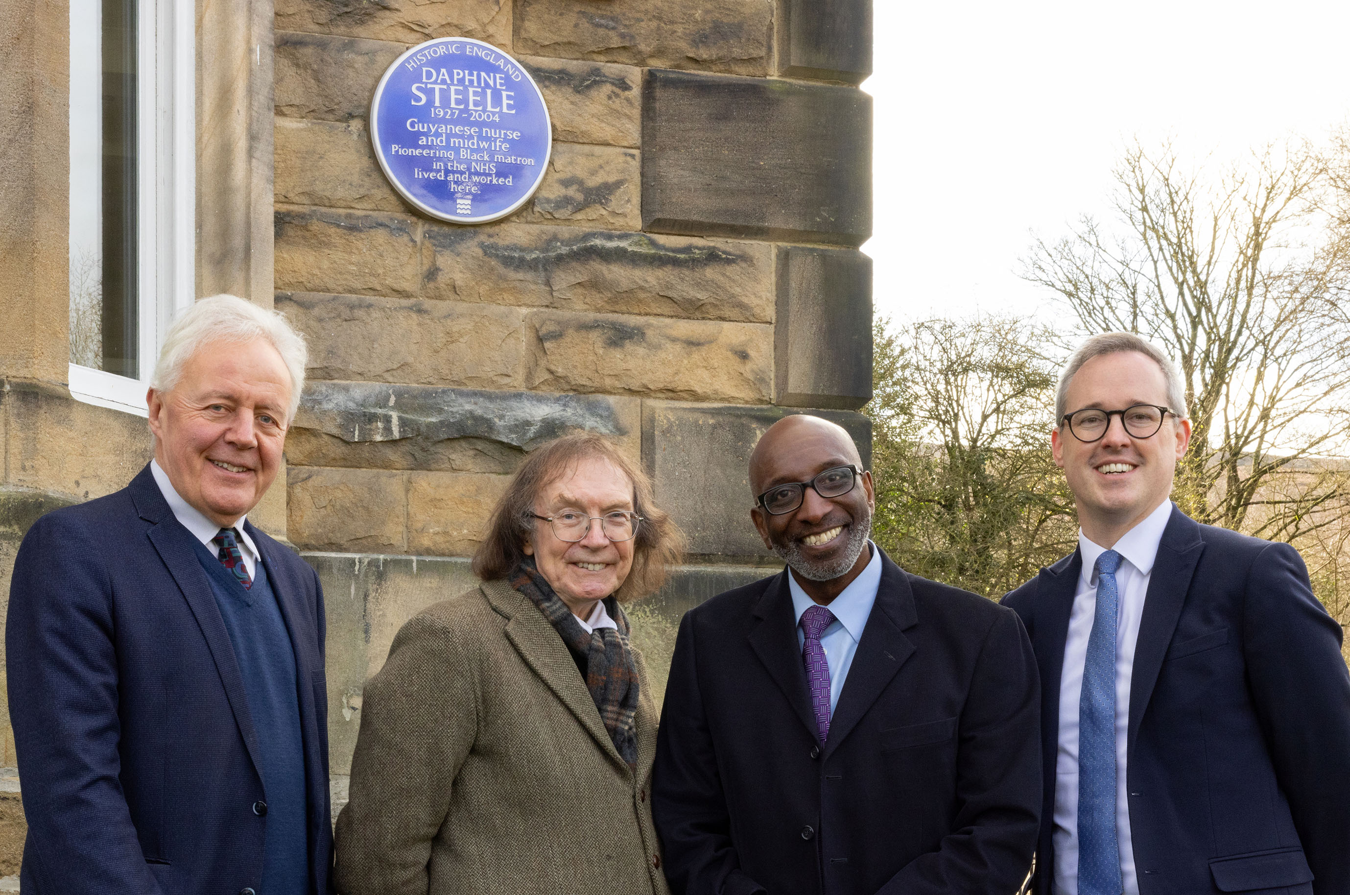 Four men in suits and coats stood smiling for the camera in the foreground, underneath a heritage blue plaque on a wall in the background.
