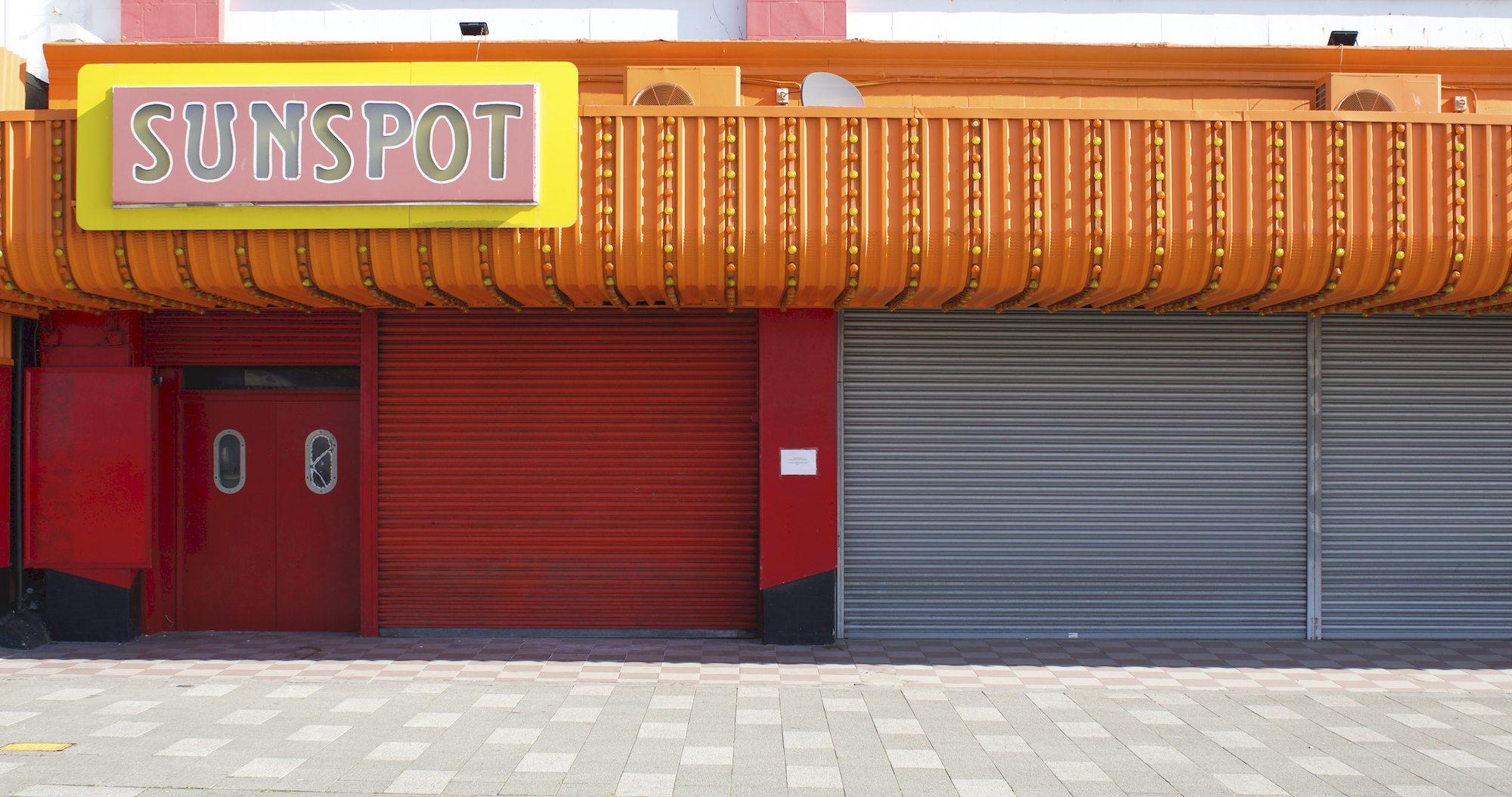 A large store front with corrugated orange facade and red shutters is closed. The shop is called 'sunspot; these words are signposted in pink and yellow. It is a warm sunny day.