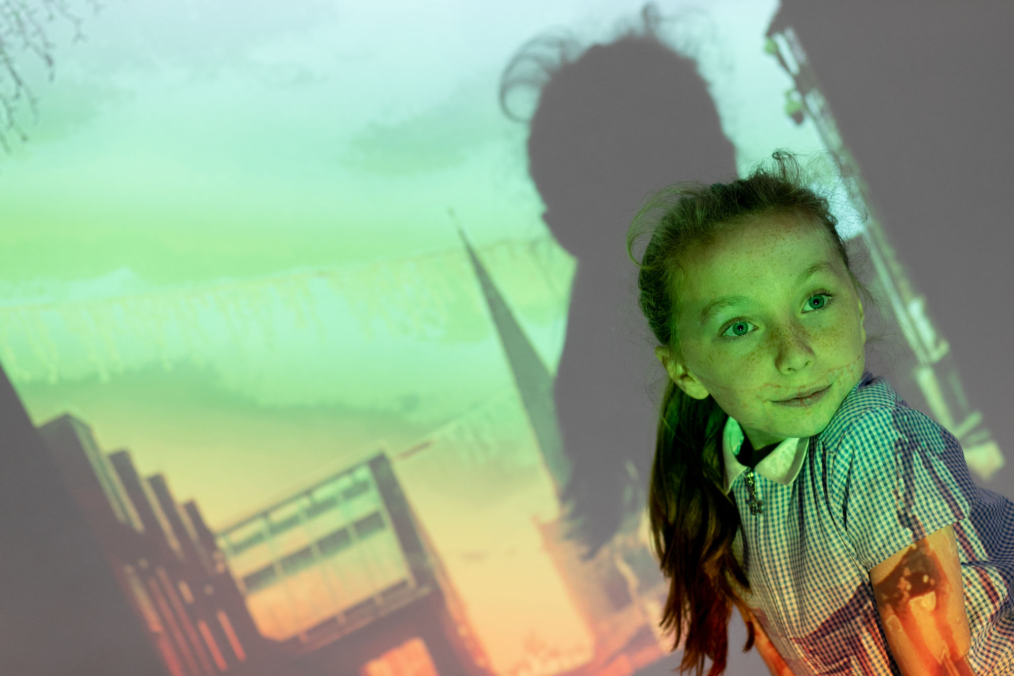 A girl in school uniform photographed in front of a photo of a street scene projected on her and a wall behind her.