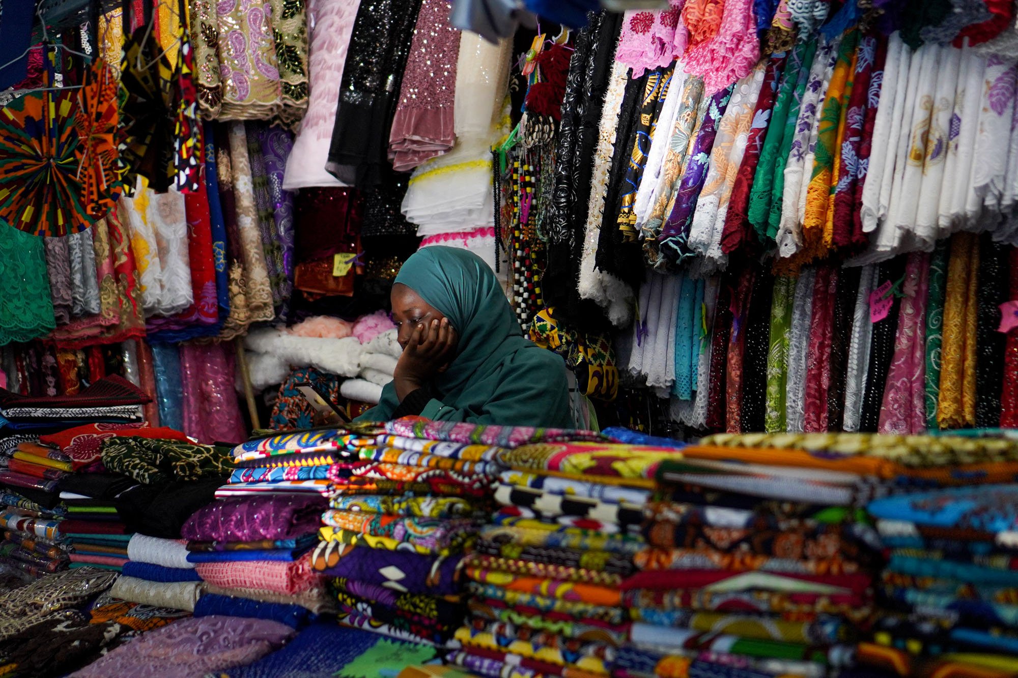 A woman props her head in her hand, leaning on piles of fabric at a market stall.