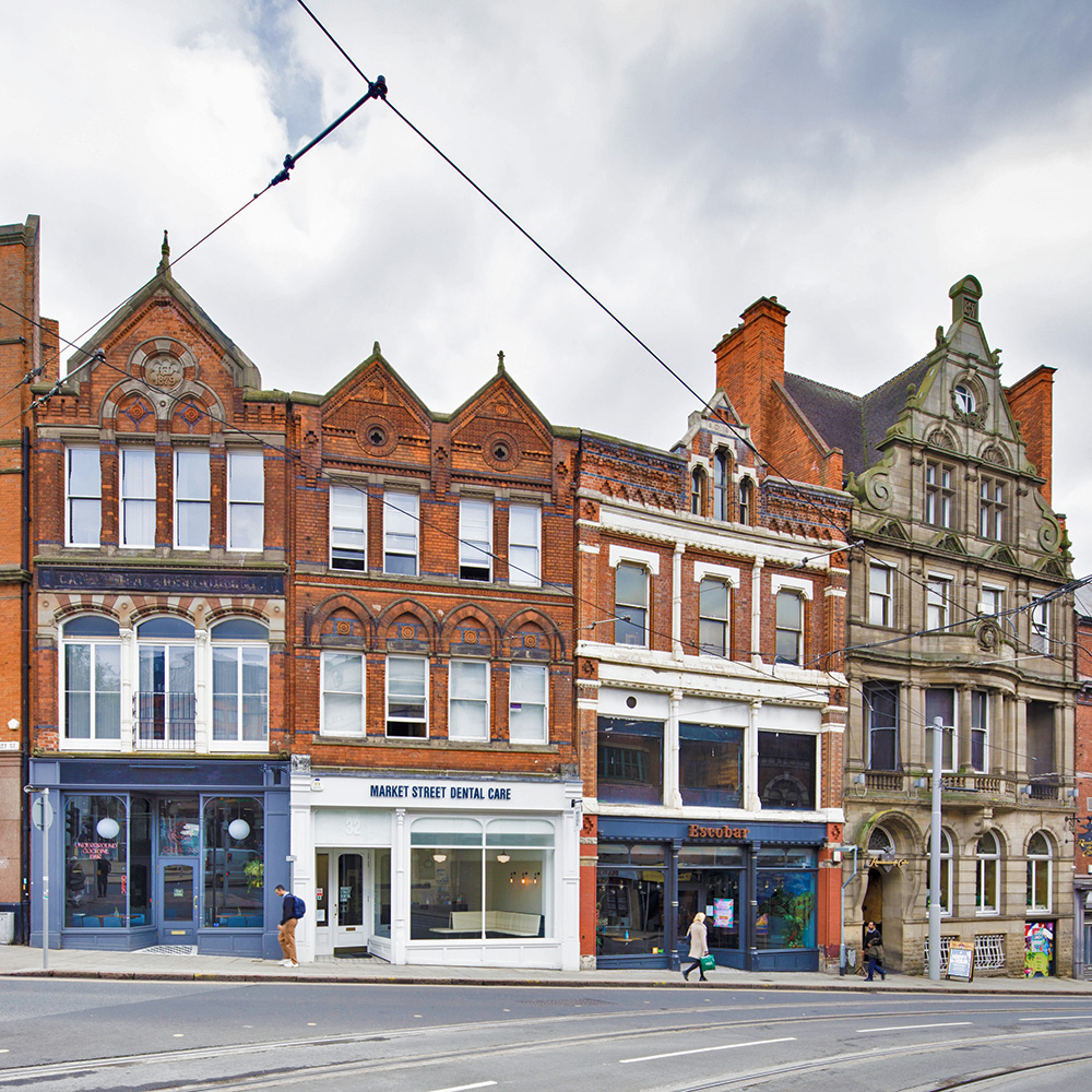 An urban high street with tall red brick shop fronts with ornate brickwork. Tram lines overhead. 