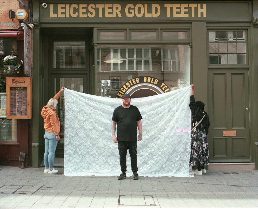 A man posing for a photo in front of the premises of Leicester Gold Teeth. Behind him, a temporary backdrop of net curtain fabric is held up on either side by two people facing away from the camera.