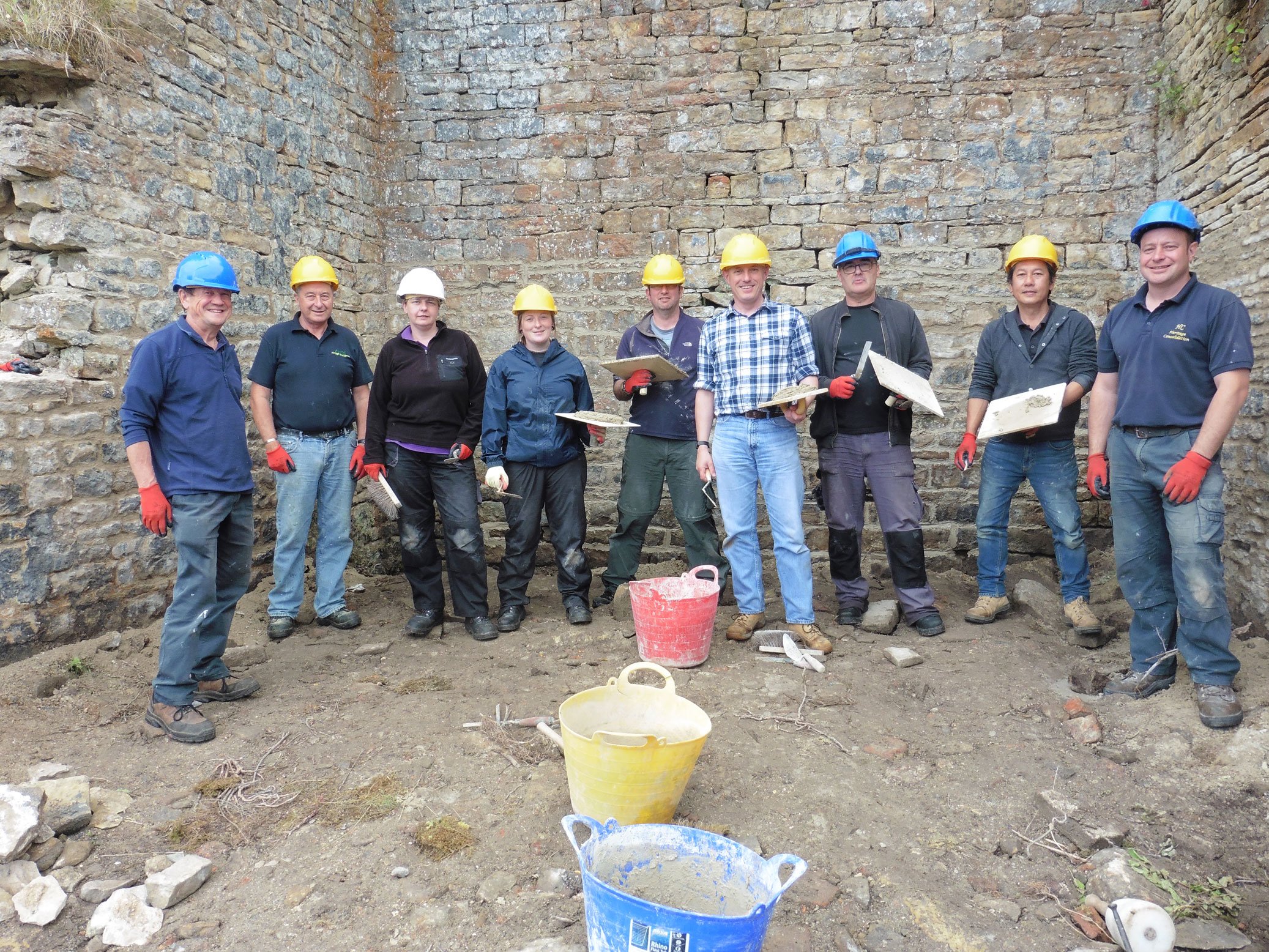  A group of volunteers with repointing tools and buckets of mortar in front of a stone building.