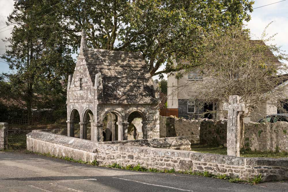 A photograph of a small, medieval stone structure enclosed by a stone wall. It has open arches around four sides. Beside it is a stone cross.