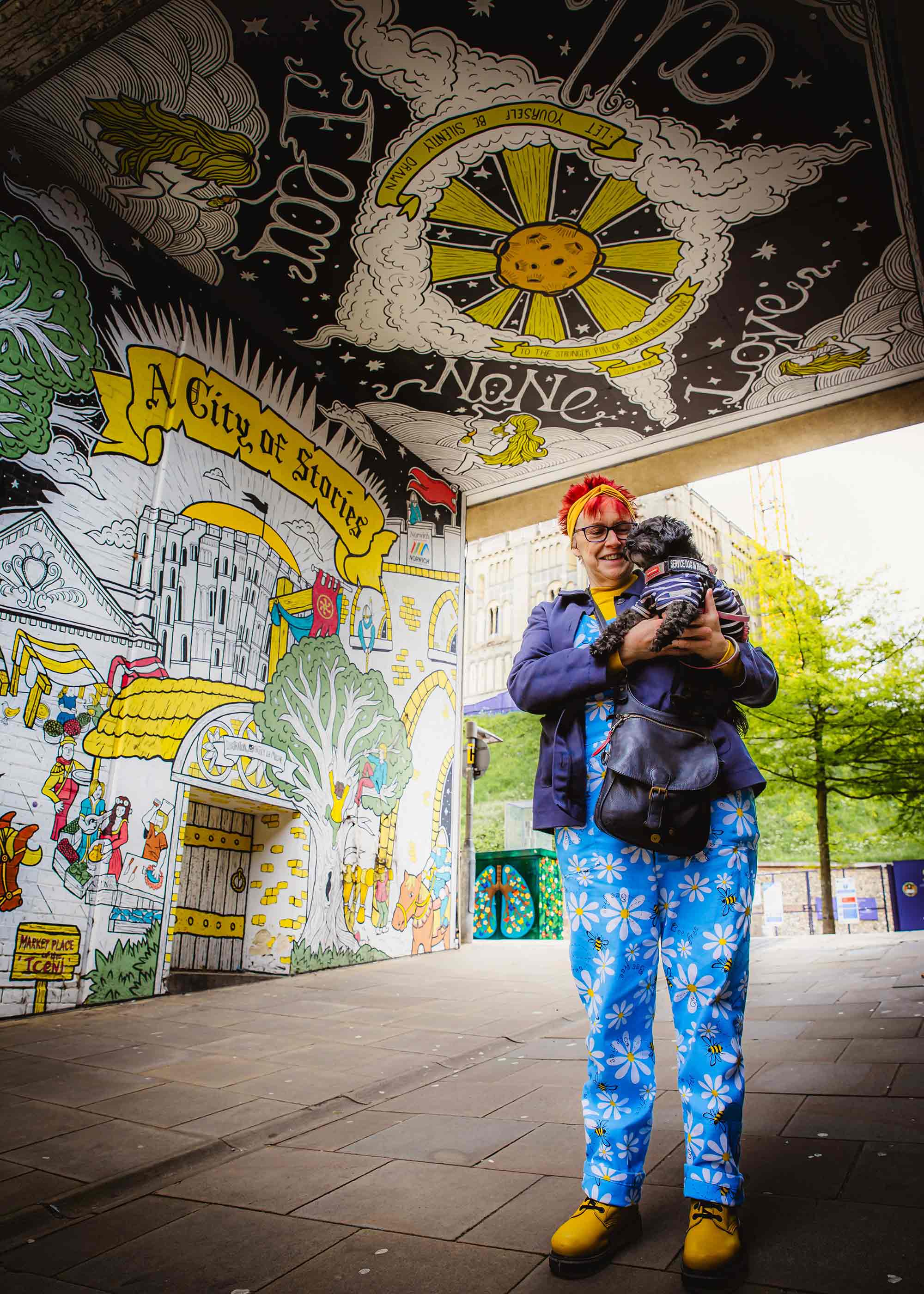 A woman holding a small dog standing under an archway which is covered with images which have been painted on it.